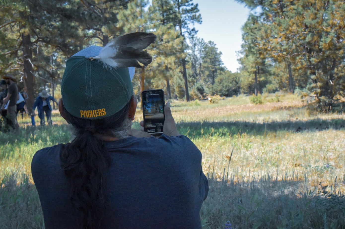 A teacher wearing a hat sits in an open forest with the GLOBE observer app, showing a tree, in front of him.