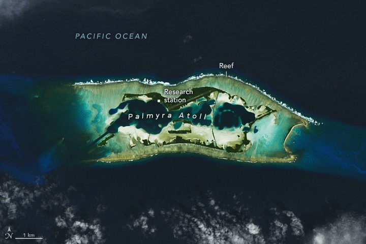 A satellite image shows a a collection of islands in various shades of green, stretching roughly in two parallel lines from left to right. On the top line, a thin layer of whitewater denotes where the reefs are. Bright teal surrounds the islands where the water is shallowest, fading quickly into the deep blue ocean water composing the majority of the image.