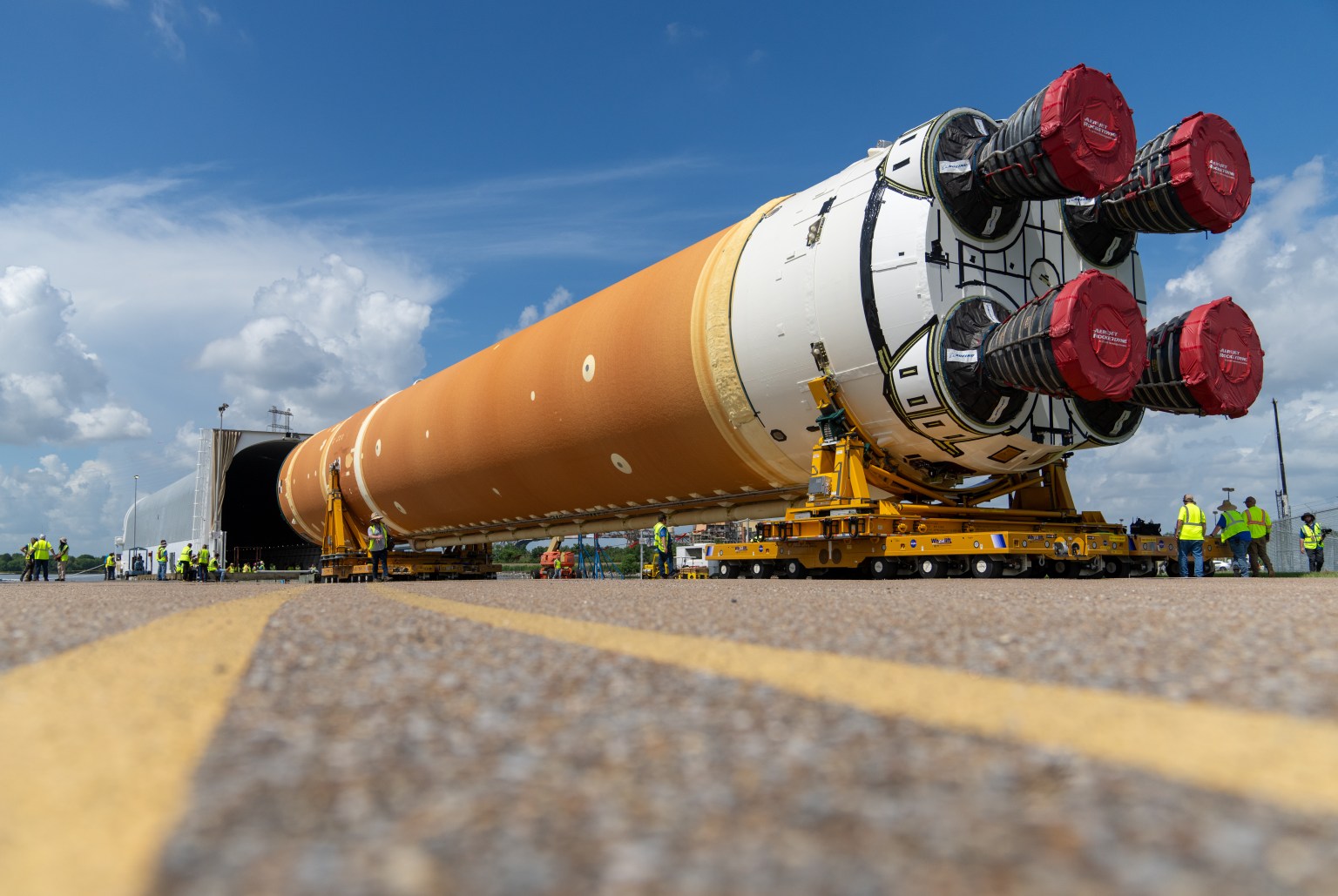 The core stage of the Artemis Space Launch System being loaded on a covered barge. The stage is a large cylinder shape with the engines facing toward the camera on two yellow transporters that are guiding the stage into a covered grey container in the background. The body of the cylinder is mostly an orange color and white around the bottom. The four engines on the bottom are covered with red material.