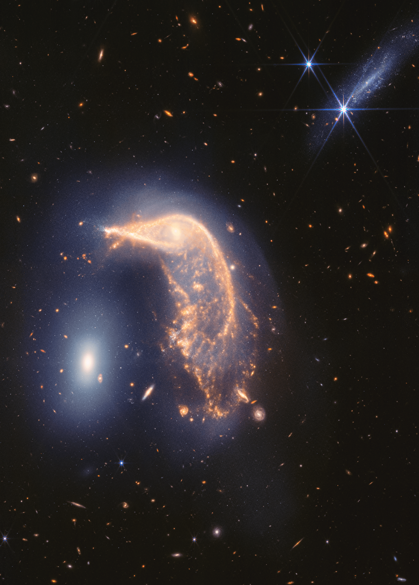 Arp 142, two interacting galaxies, observed in near- and mid-infrared light. At left is NGC 2937, nicknamed the Egg. Its center is the brighter and whiter. There are six diffraction spikes atop its gauzy blue layers. At right is NGC 2936, nicknamed the Penguin. Its beak-like region points toward and above the Egg. Where the eye would be is a small, opaque yellow spiral. The Penguin’s distorted arms form the bird’s beak, back, and tail. The tail is wide and layered, like a beta fish’s tail. A semi-transparent blue hue traces the Penguin and extends from the galaxy, creating an upside-down U over top of both galaxies. At top right is another galaxy seen from the side, pointing roughly at a 45-degree angle. It is largely light blue. Its length appears approximately as long as the Egg’s height. One foreground star with large, bright blue diffraction spikes appears over top of the galaxy and another near it. The entire black background is filled with tiny, extremely distant galaxies.