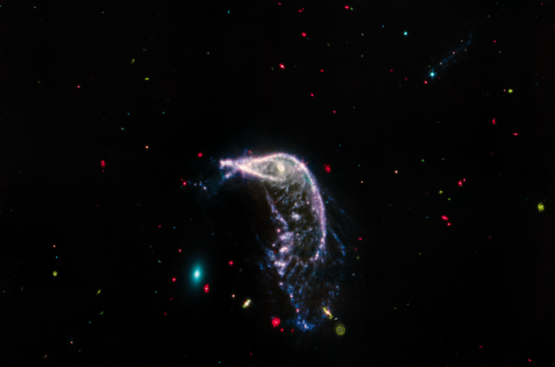 Two interacting galaxies known as Arp 142 in a horizontal image taken in mid-infrared light. At left is NGC 2937, an elliptical galaxy that looks like a tiny teal oval and is nicknamed the Egg. At right is NGC 2936, a distorted spiral galaxy nicknamed the Penguin, which is significantly larger. A beak-like region points toward the Egg, but lies far above it. Where the eye would be is an opaque, almost washed-out pink spiral. This galaxy’s distorted pink, purple, and blue arms create the bird’s beak, back, and tail. The tail, which is closer to the Egg, is wide and layered, like a beta fish’s tail. The Penguin and the Egg appear very separate. The galaxy at top right, PGC 1237172, is barely visible. A brighter slightly larger blue foreground star that is overtop this galaxy has tiny diffraction spikes. Throughout the image are tiny galaxies in bright reds, greens, and blues. The background of space is black.