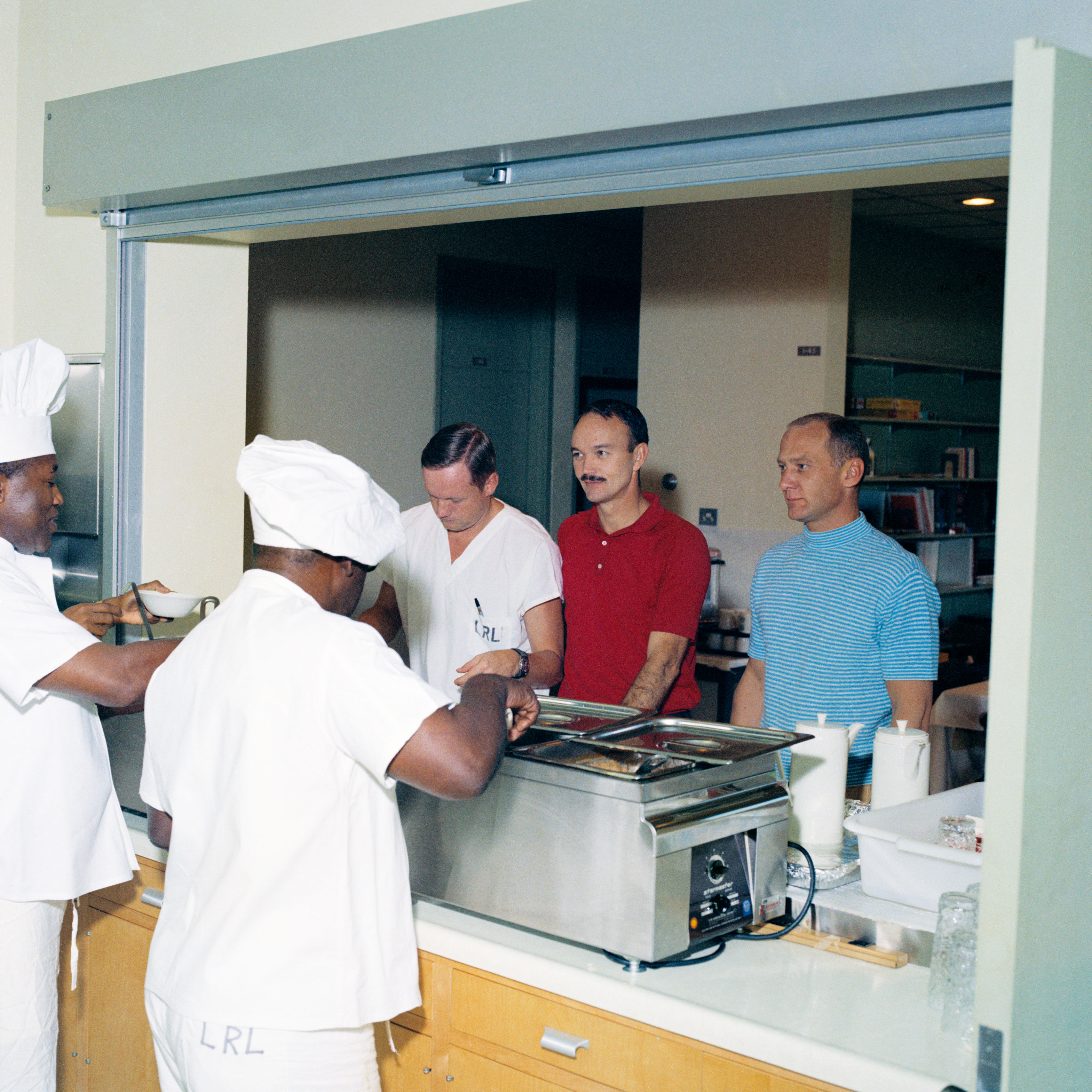 In the Lunar Receiving Laboratory (LRL) at the Manned Spacecraft Center, now NASA’s Johnson Space Center in Houston, Apollo 11 astronauts Neil A. Armstrong, left, Michael Collins, and Edwin E. “Buzz” Aldrin line up for food in the LRL’s dining area