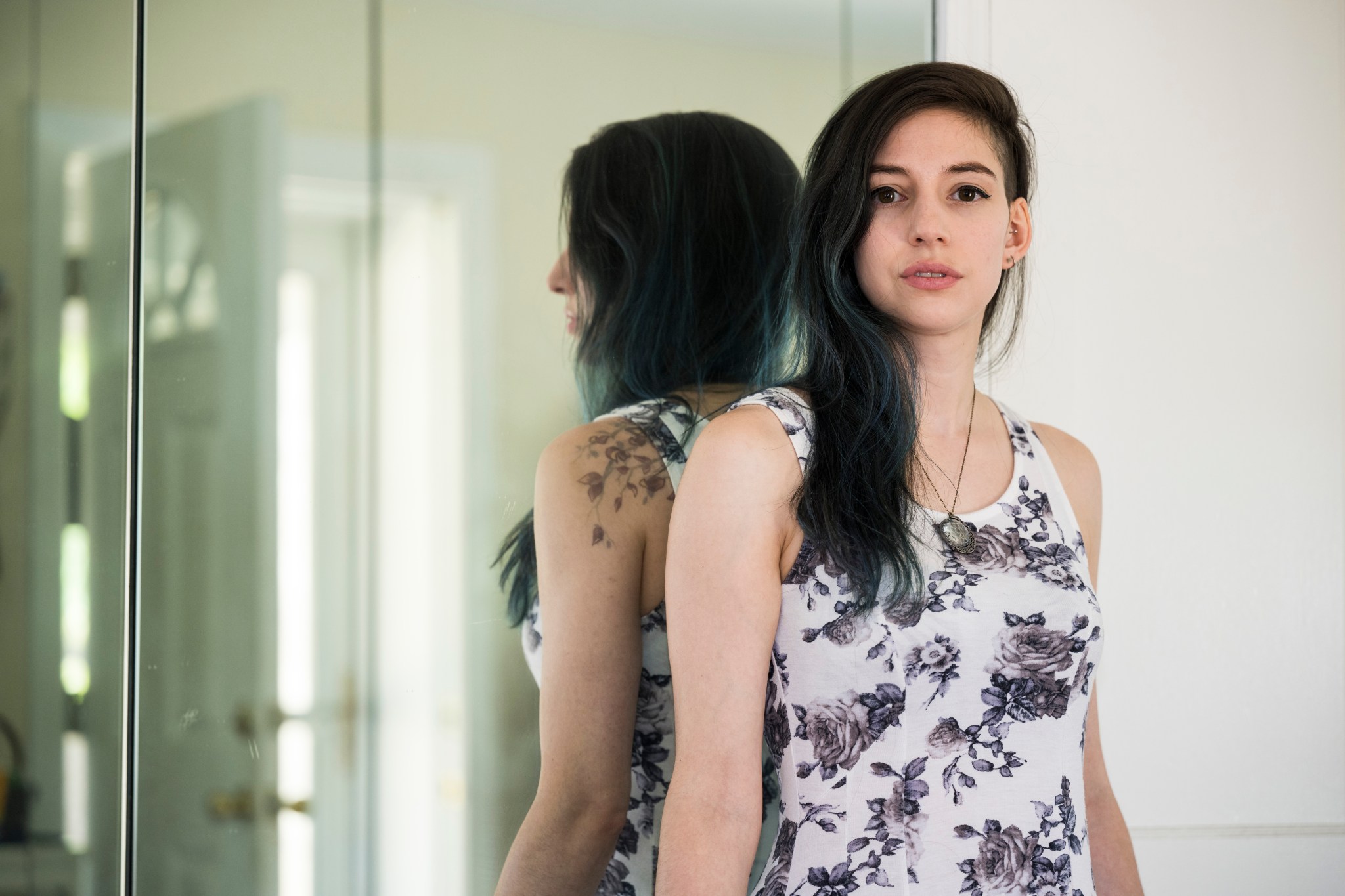 A woman with long, dark brown hair and subtle blue highlights stares head on at the camera wearing a white tank top with blue and purple roses on it.