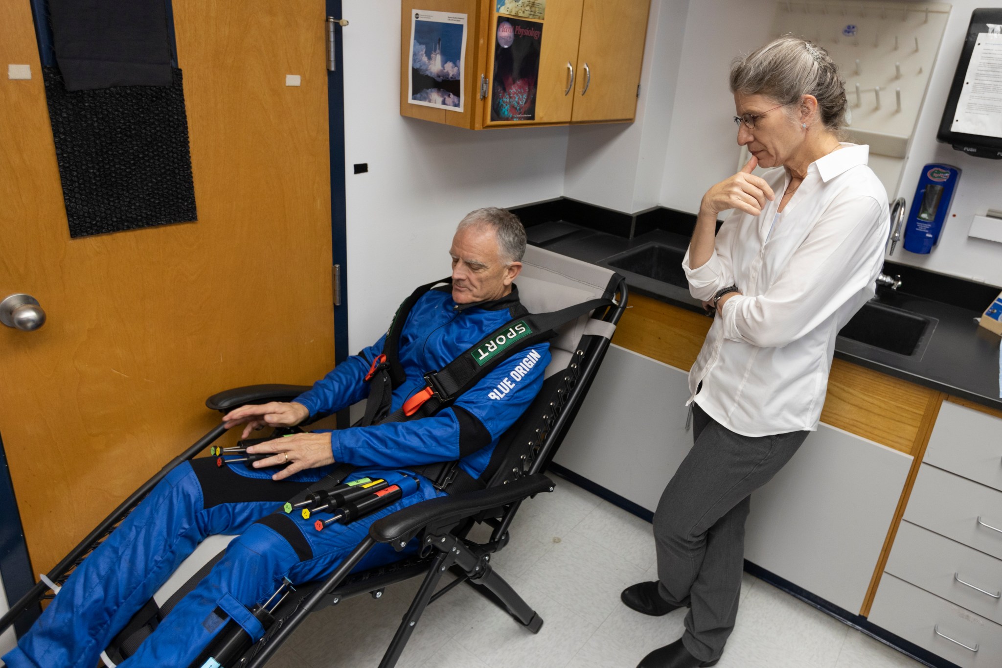 First NASA-Supported Researcher to Fly on Suborbital Rocket in reclined chair handles tubes attached to his thighs while woman watches.