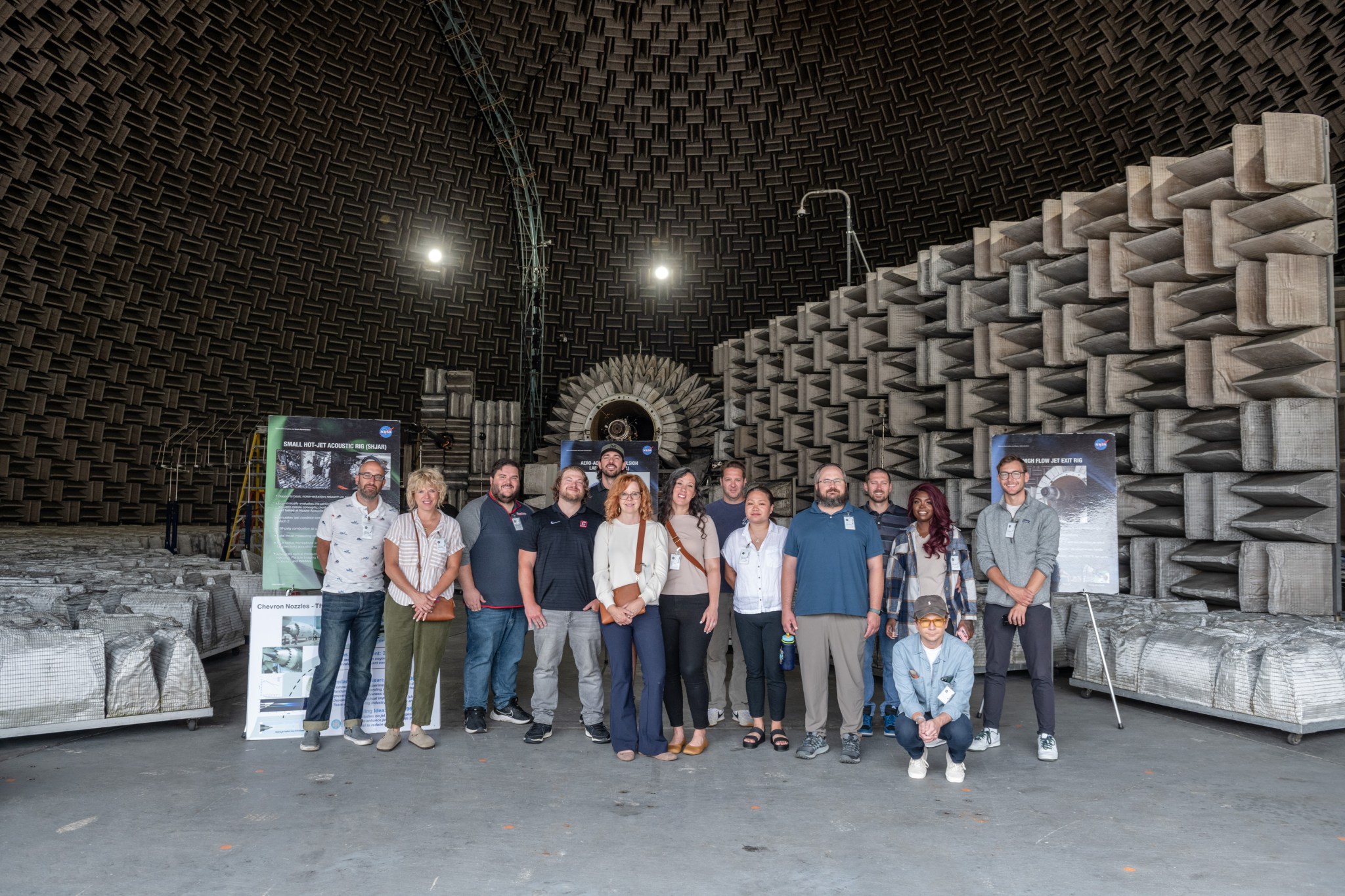 A large group of people stand in a large laboratory with fiberglass wedges mounted on the dome’s interior walls and floor areas adjacent to the test rigs.