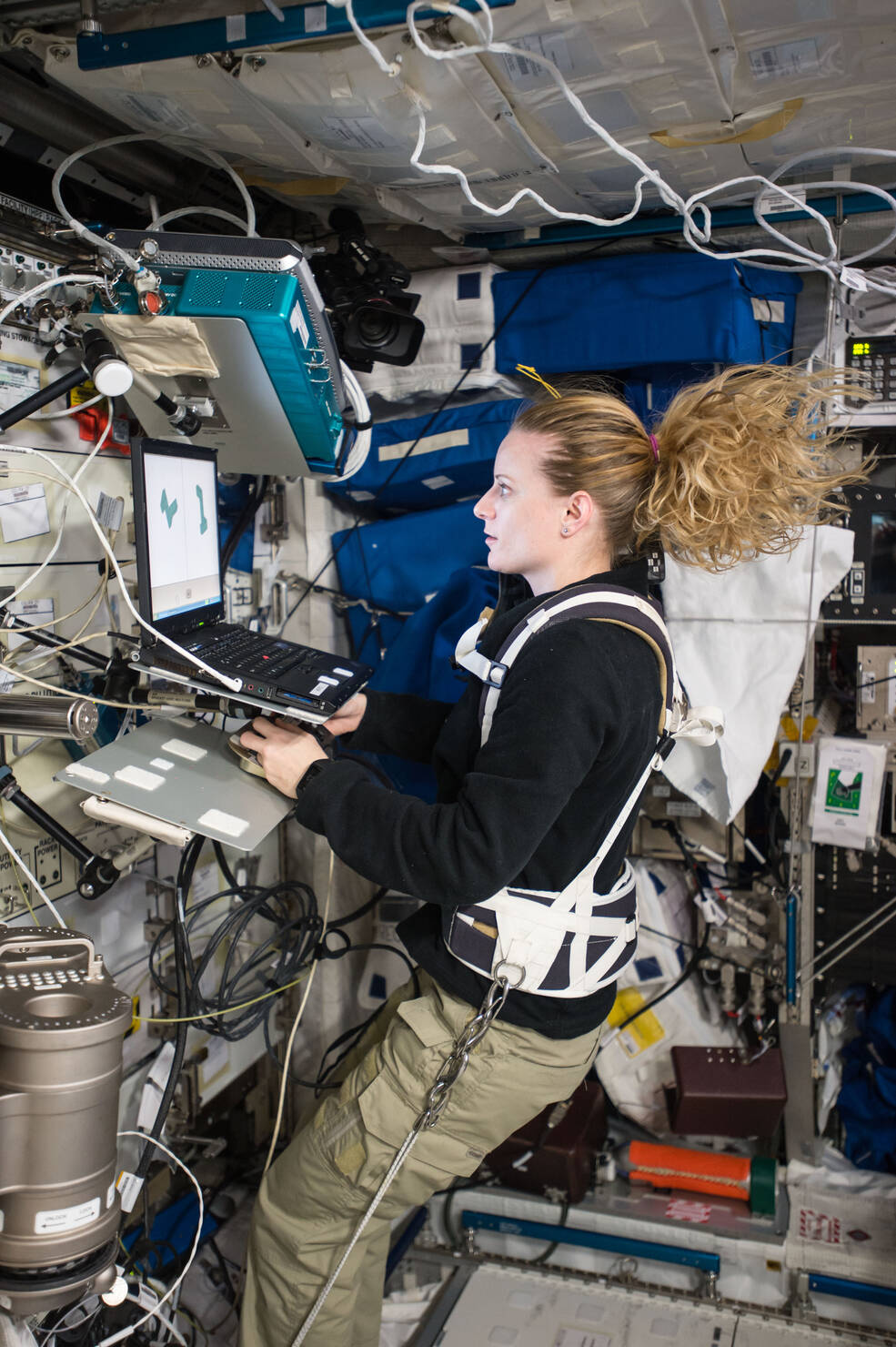 Rubins, wearing a black shirt and khaki pants, with her hair in a ponytail floating above her head and a harness around her upper body that tethers her to the surface beneath her, works a controller in front of a laptop. There are blue storage bags behind her and other equipment and cords on the wall in front of her.