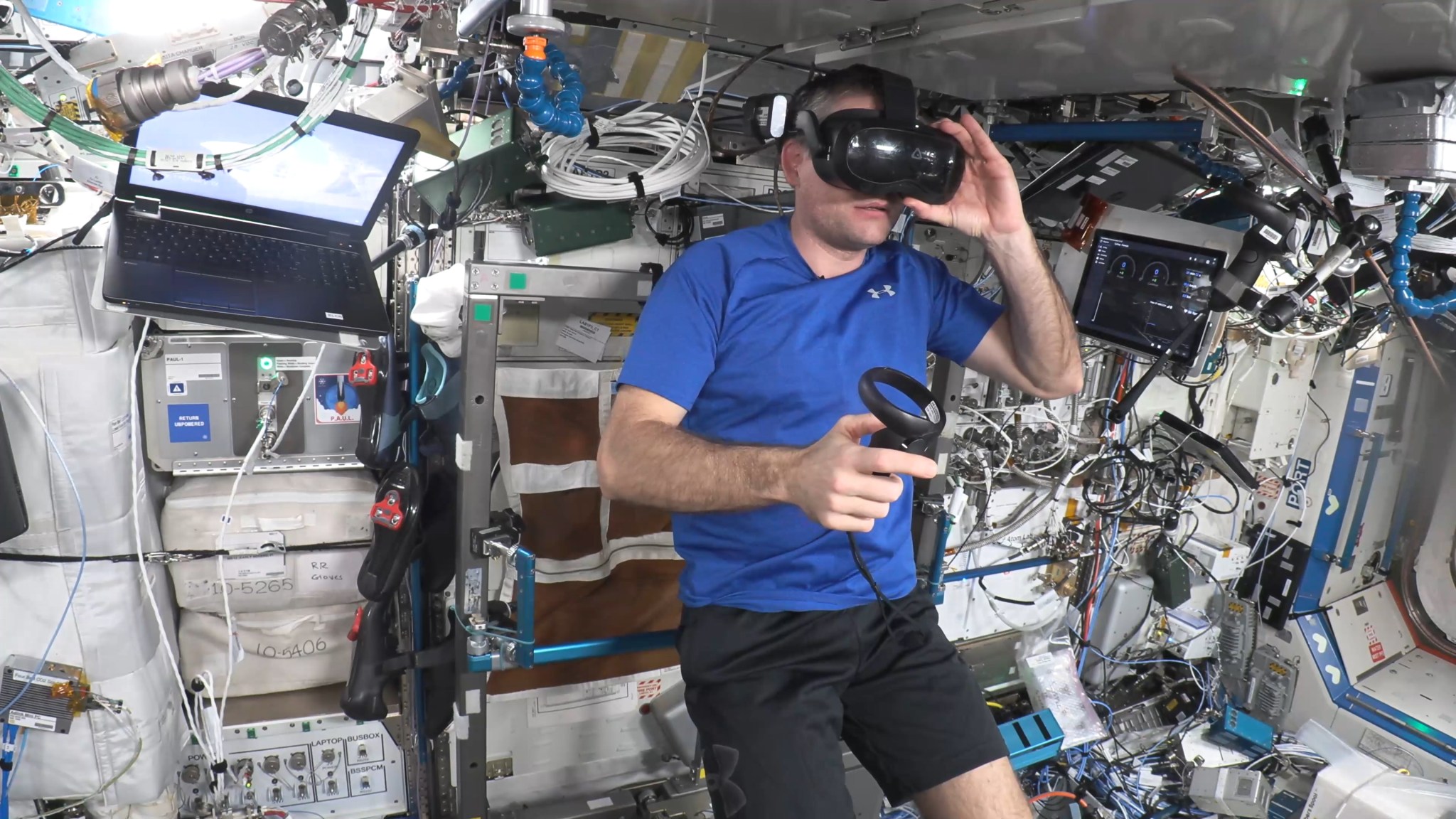 Mogenson, in a blue t-shirt and black shorts, is wearing a black VR headset and adjusting it with his left hand and holding a controller in his right hand. There is a laptop screen on either side of him and multiple cords and cables on the station wall behind him.