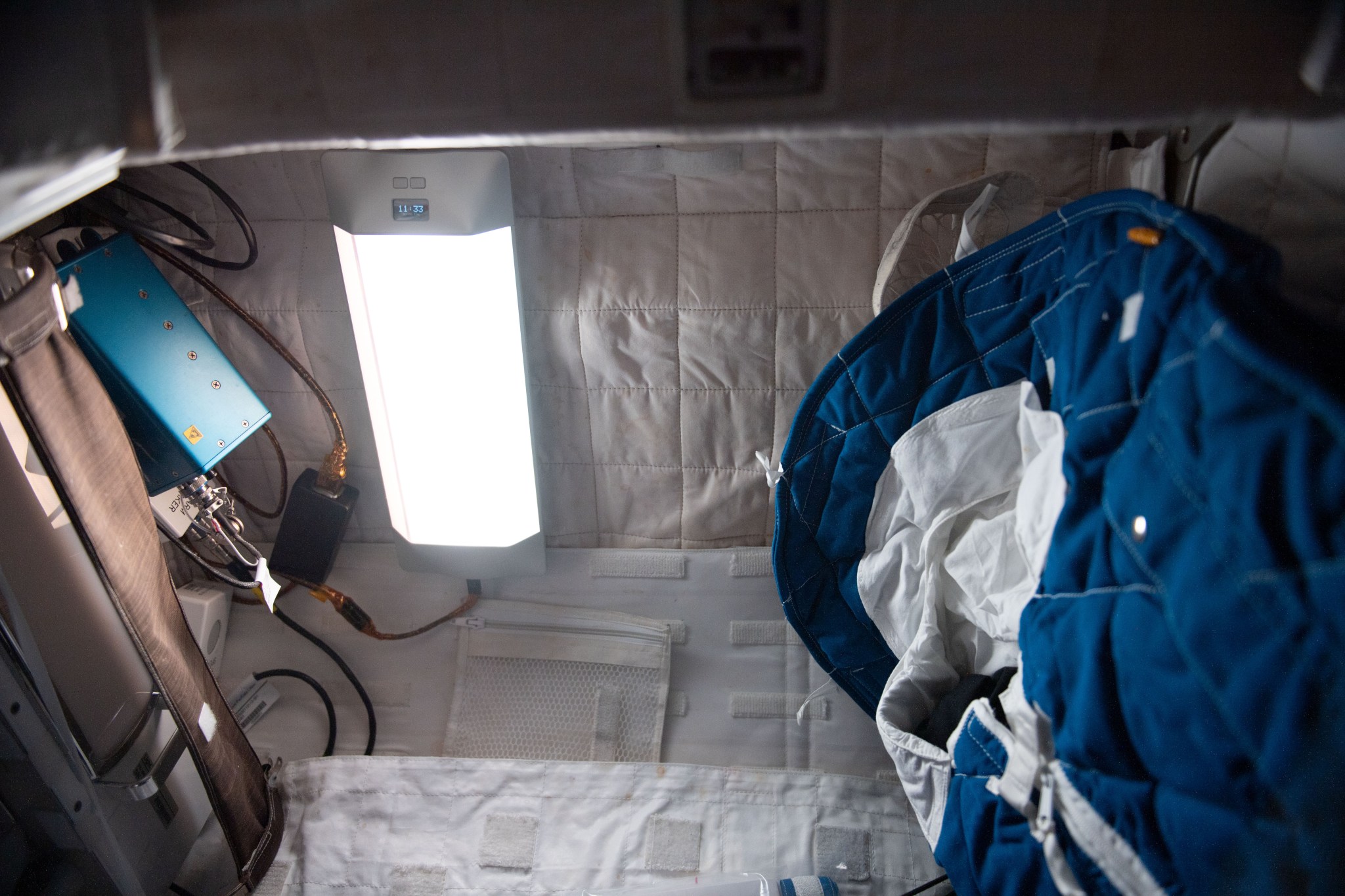 A rectangular white light about the size of a computer monitor is attached to the quilt-like ceiling at the top of this image. A blue and white sleeping bag is visible on the right of the image and on the left, a blue brick-sized power box is connected to the light with a cord.