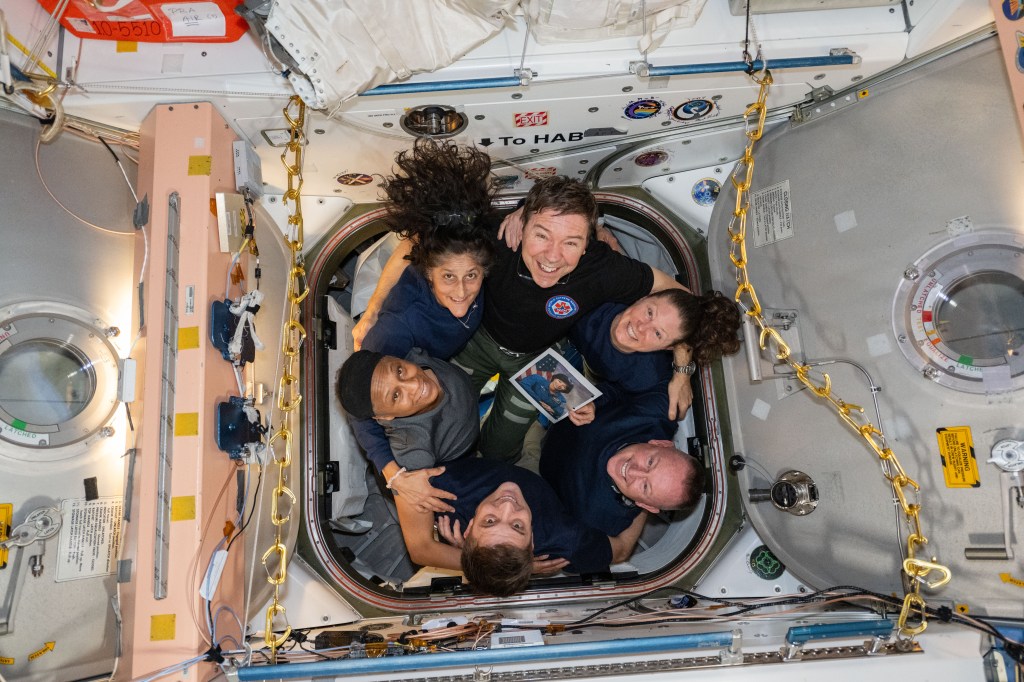 Clockwise from bottom, NASA astronauts Matthew Dominick, Jeanette Epps, Suni Williams, Mike Barratt, Tracy C. Dyson, and Butch Wilmore, pose for a team portrait inside the vestibule between the Unity module and the Cygnus space freighter from Northrop Grumman. Dyson holds a photograph of NASA astronaut Patrica Hilliard for whom the Cygnus spacecraft, S.S. Patricia “Patty” Hilliard Robertson, is named after.