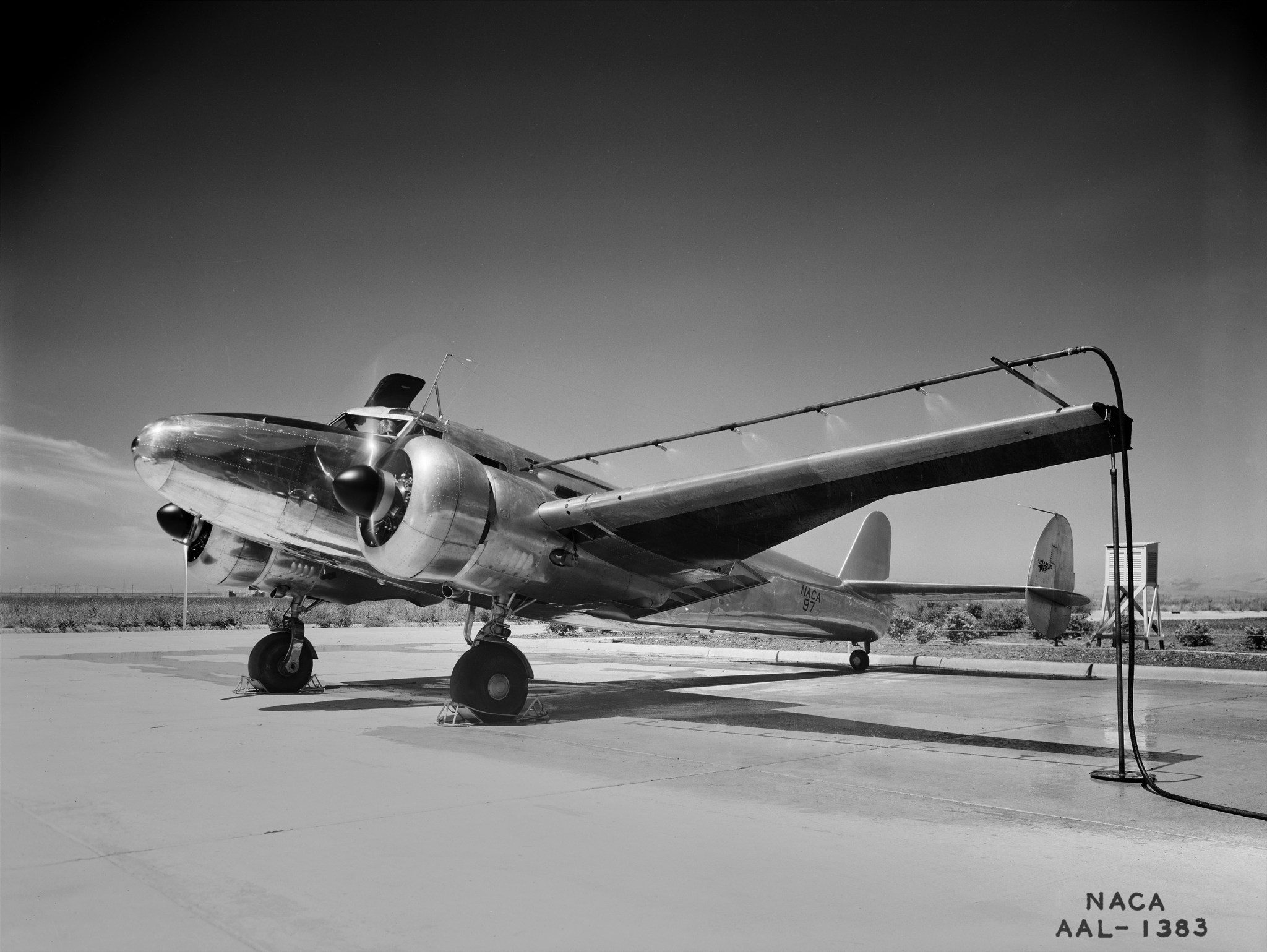 A black and white photo of a Lockheed 12A airplane sitting on a tarmac, with pipes installed along its wings that are blasting warm air on the leading edge of the wing, studying aviation de-icing.