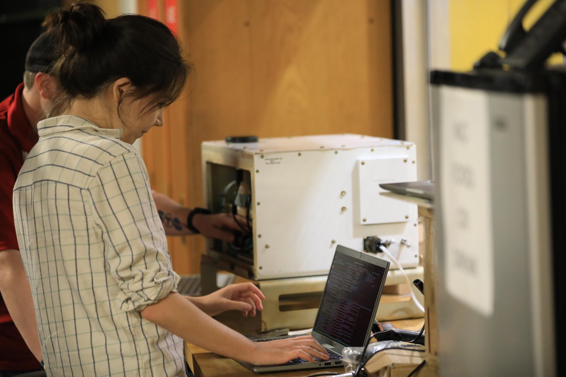 A woman with brown hair pulled into a bun, wearing a white, collared shirt with black lines, stands in the foreground of the photo. She is working on a grey laptop computer with black screen with computer coding shown. Behind her, on the left side, is the side of a man’s head and he is wearing a red polo. On the right side, behind her computer, is a white cube with wires and the man is placing his hand inside.