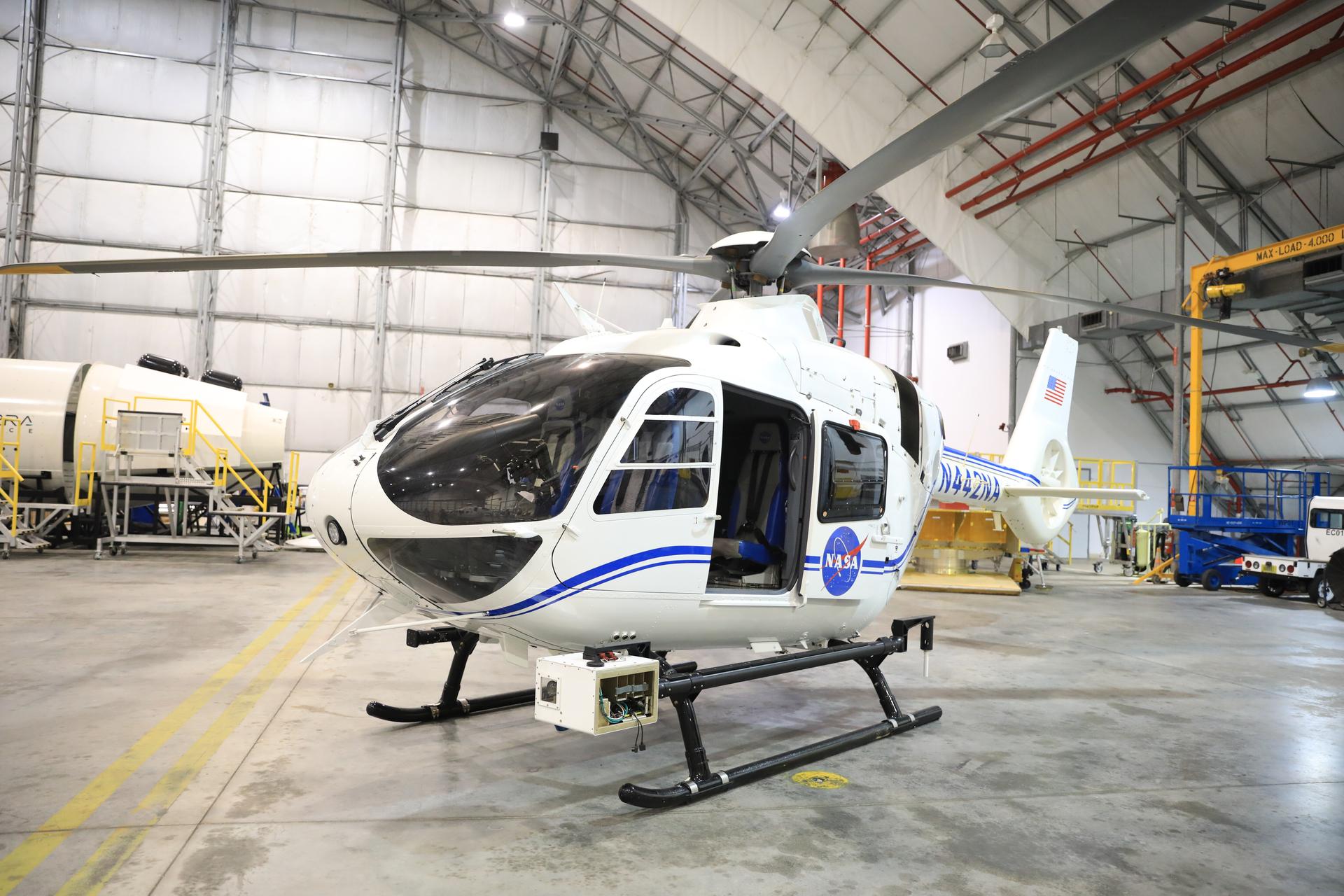 A white helicopter with blue stripe and NASA logo sits inside of an aircraft hangar with grey cement floors and white roofing with metal beams. The helicopter has four grey blades and has a black base. A white cube is attached to the black base and holds wires and cameras. No one sits inside the helicopter, but the door is open, and a grey seat is shown along with four black, tinted windows. There is an American flag on the helicopter’s tail.