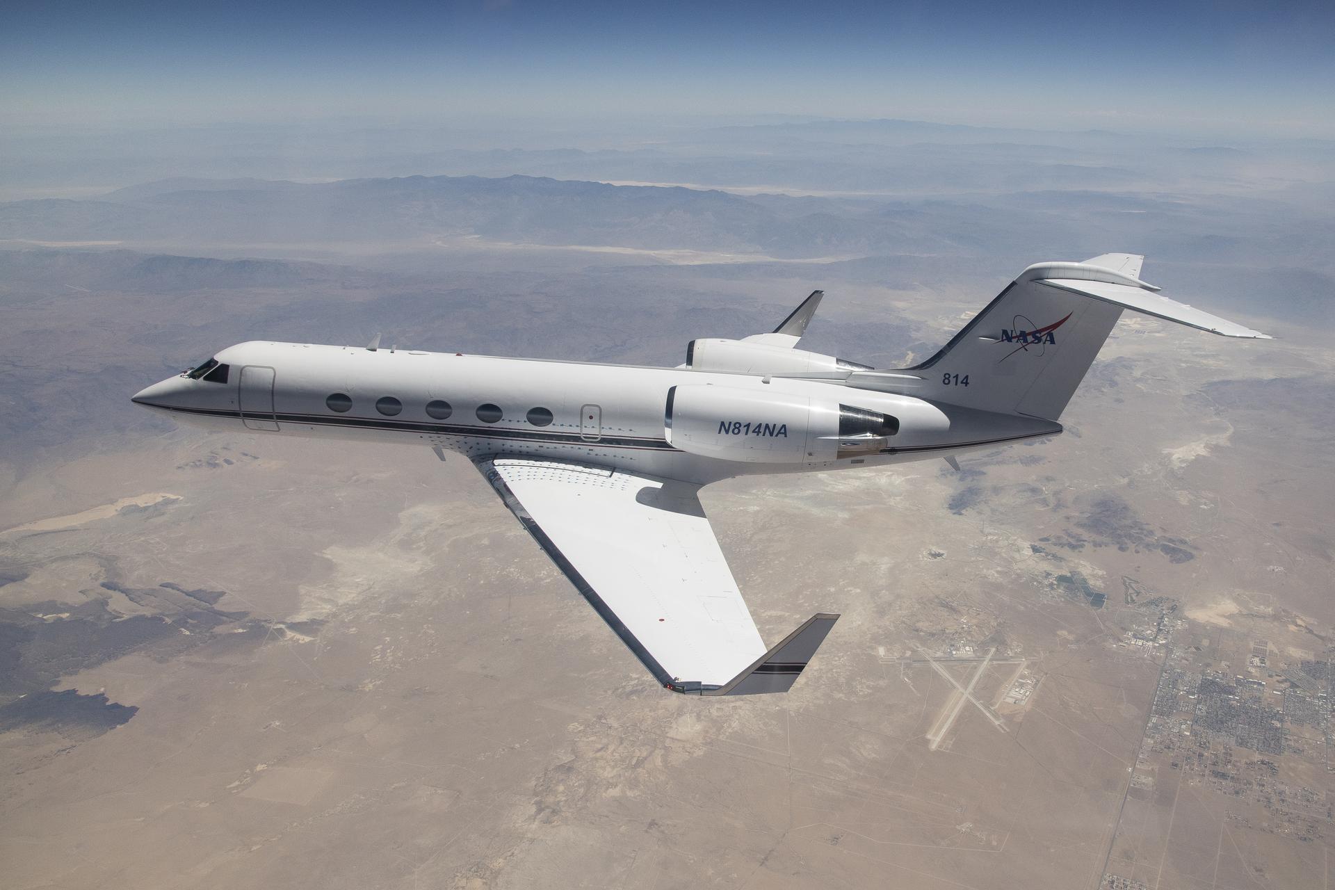 A white Gulfstream IV airplane flies to the left of the frame over a tan desert landscape below and blue mountain ranges in the back of the image. The plane’s tail features the NASA logo, and its wings have winglets. Visible in the lower right third of the image, directly behind the airplane’s wingtip is the Mojave Air and Space Port in Mojave, California. 