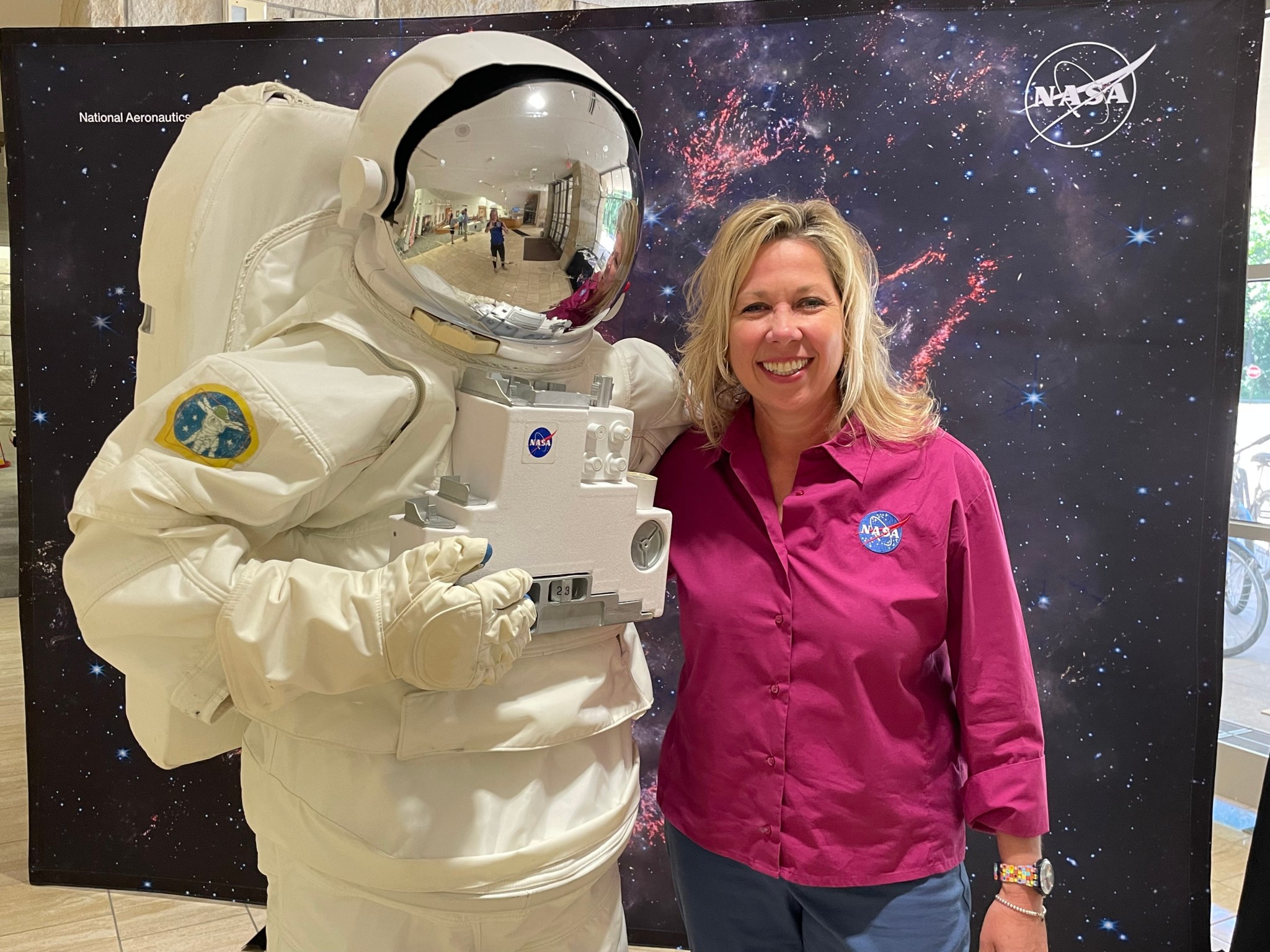 An astronaut mascot poses with a member of NASA’s Transportation Integration Office in front of a black backdrop with pink and blue stars.