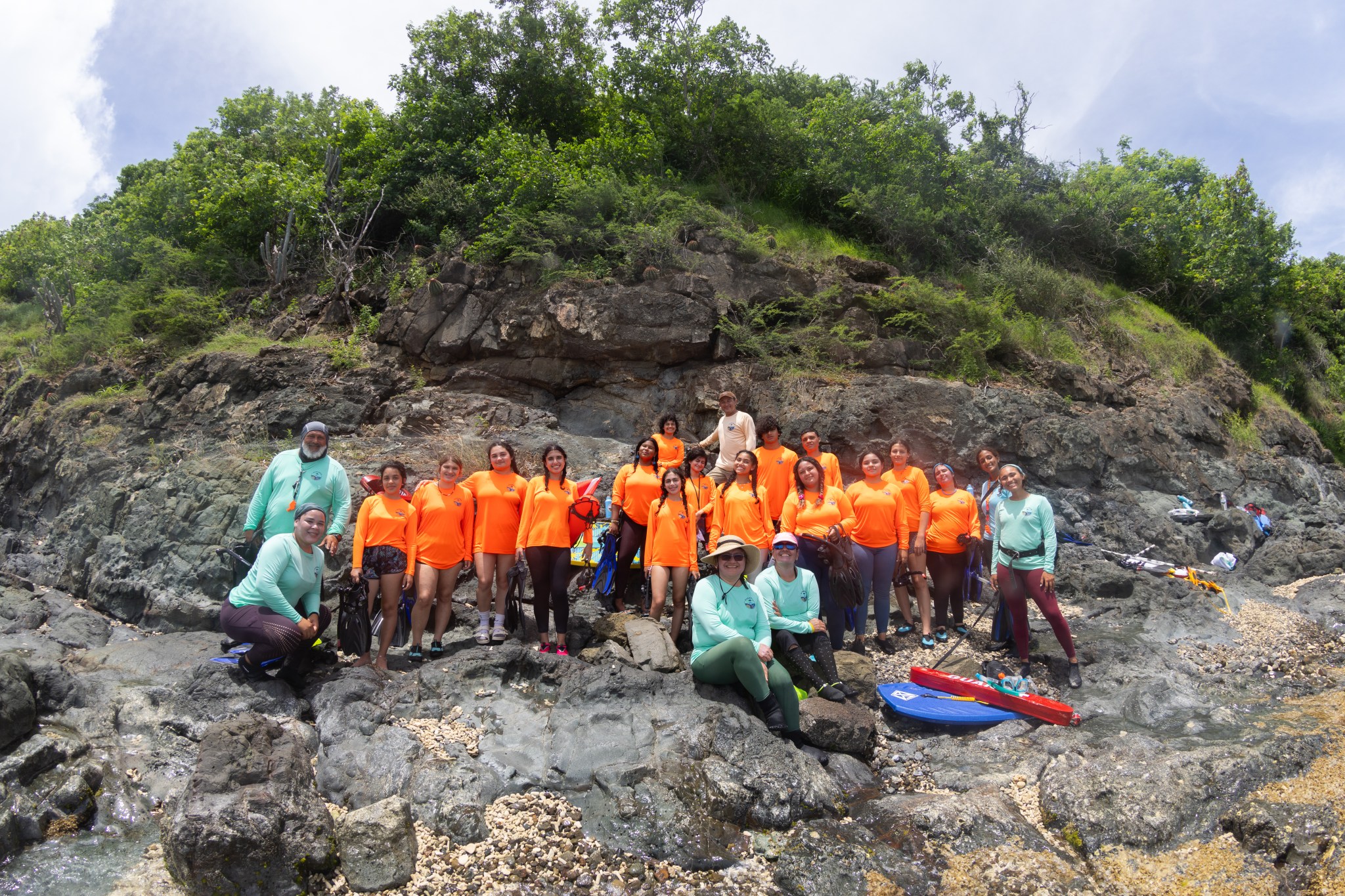 Fifteen high schoolers in bright orange long-sleeve shirts stand on dark grey rock littered with tan sand. Behind them is a small cliff of the same rock, topped in bright green foliage. On either side and sitting in front of the students are five instructors, wearing teal long-sleeve shirts.