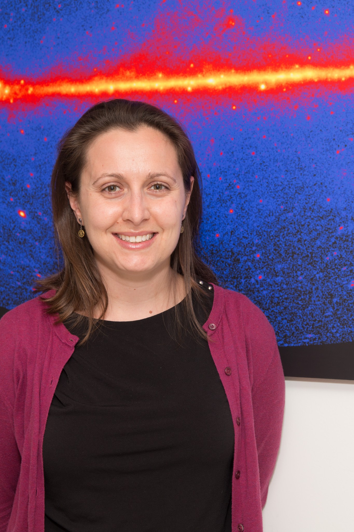 Regina Caputo stands in front of a white wall with an image hanging on it. She is smiling and wearing a black shirt under an open maroon cardigan. Her brown hair falls loosely on her shoulders. The image behind Regina consists of a black background and in front of it a large blue oval that is not completely visible within the screen. The blue oval takes up most of the image and is speckled with deeper blues as well as some red dots. Just above Regina’s head, within the blue oval, is a horizontal orange and yellow line that glows. Scattered around the line are a few glowing yellow dots. Additionally, red speckling surrounds the line. The speckling starts dense towards the orange and yellow line — dense enough to appear solid — and becomes less dense further from the center line.