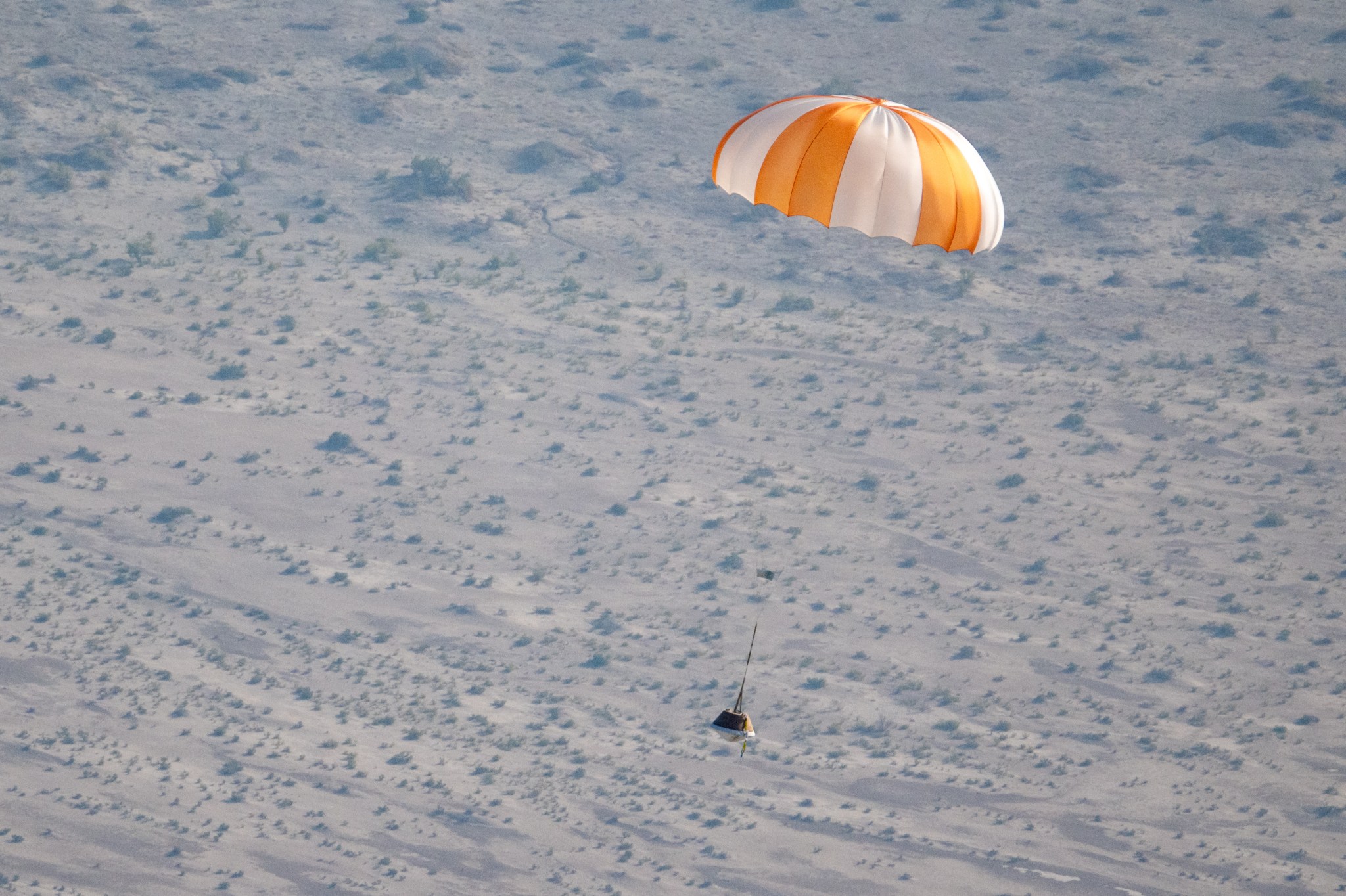 An orange and white parachute is visible at top right. It is attached to a training model of a capsule. The black and white cone-like capsule is quite small in this image. The pale ground below makes up the background of the image.