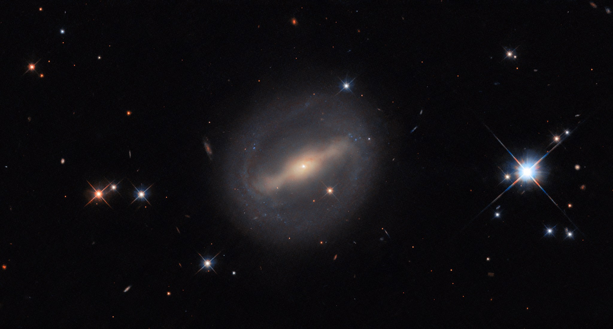 An almost circular galaxy is at the center of the image. It has a glowing bar stretching across its core; from the ends of the bar, thin spiral arms wrap around the galaxy to form a closed disk. The arms are fuzzy from the dust and stars they contain. The galaxy is on a black, mostly empty background. A few foreground stars with cross-shaped diffraction spikes can be seen, as well as some distant galaxies in the background.