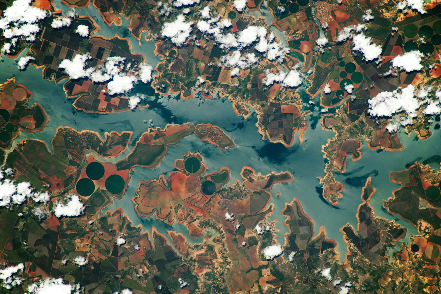 A multi-colored area of land is split horizontally by a cloudy blue river. The river cuts into the land at many points. The land is various shades of green, red, and yellow. Clumps of white clouds dot the top part and a little bit of the bottom land.