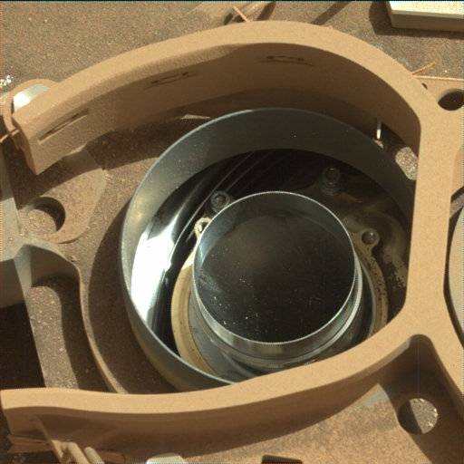 A close-up, overhead photo of a part of the Curiosity rover shows an enclosure that looks like a backwards “C.” Inside that is a circular, silver metal opening that looks like the wall of a springform cake pan, and inside that is a round opening that looks like a metal duct pipe, the inside of which is dark and you can’t see the bottom. The metal parts appear shiny and clean, but everything else in the image appears covered in brownish-orange dust.