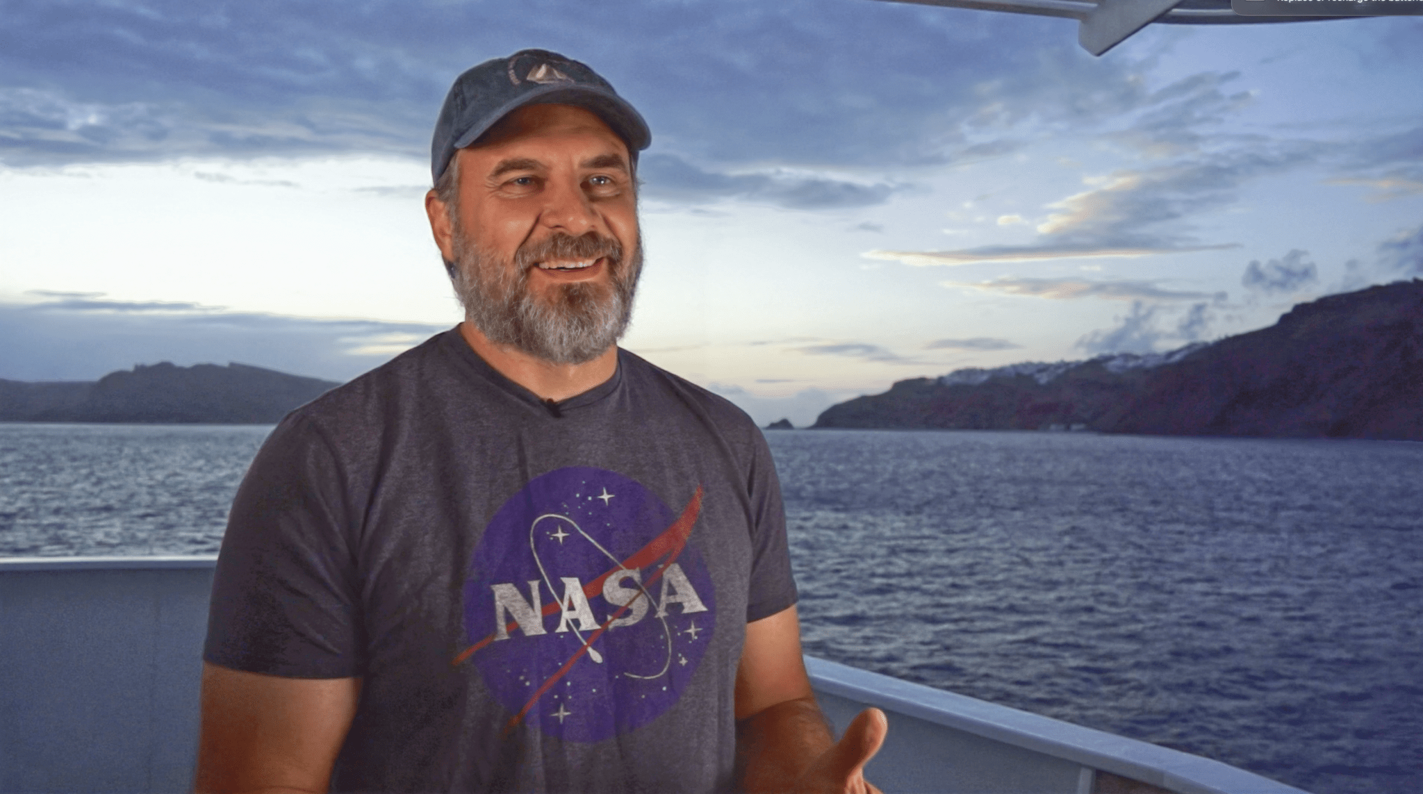 A middle-aged white man with a grayish-blonde beard smiles and stands on the deck of a ship, sun setting behind him. He is wearing a hat with a sailboat on it, and a gray NASA shirt.