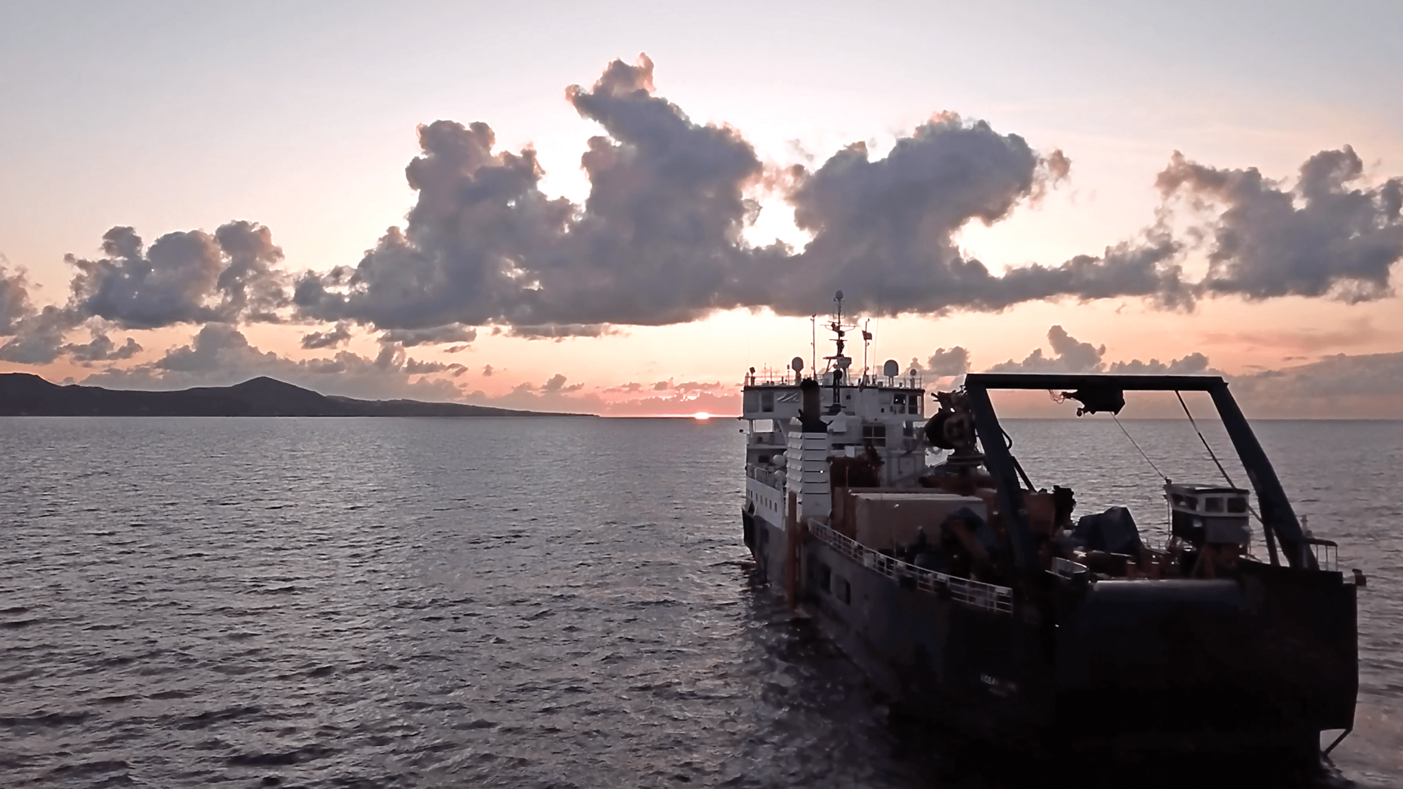 A large research ship in the lower right corner is making its way towards the horizon, as the sun sets casting rose-colored light across clouds in the sky. On the left of the image, an island is in the distance.