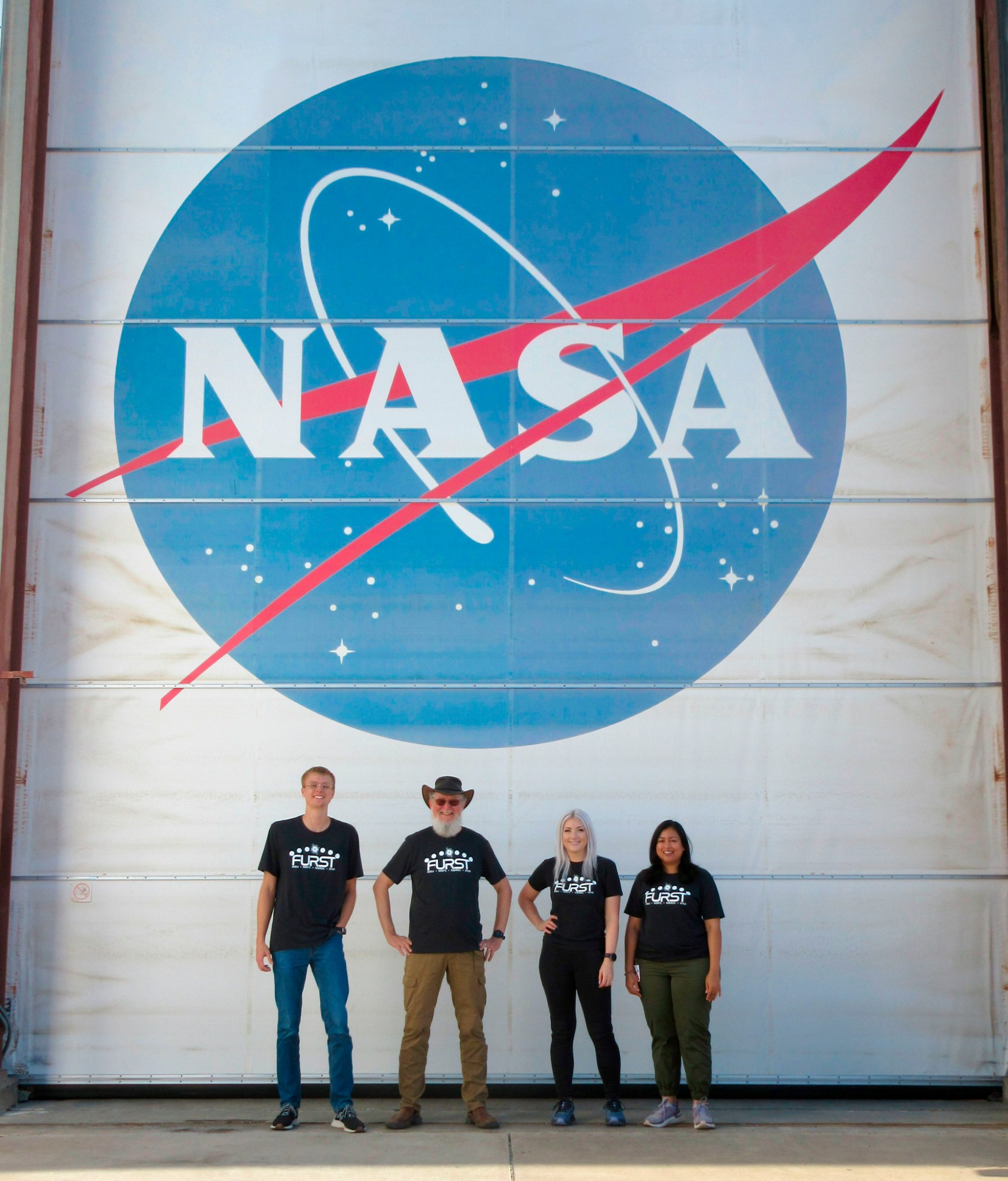 Four men stand under a large NASA meatball logo that is painted on a wall.