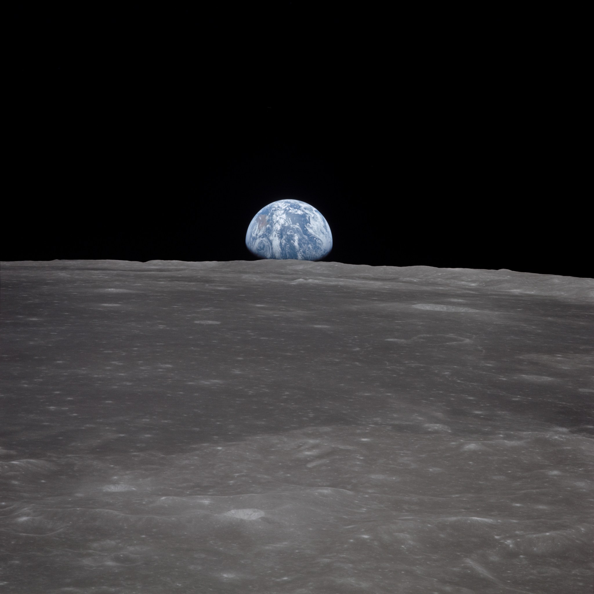 In the foreground, a grey surface stretches halfway up the photo. A small, blue and white sphere is peaking over the horizon of the grey surface, suspended against a black sky. The lower part of the sphere is shadowed.