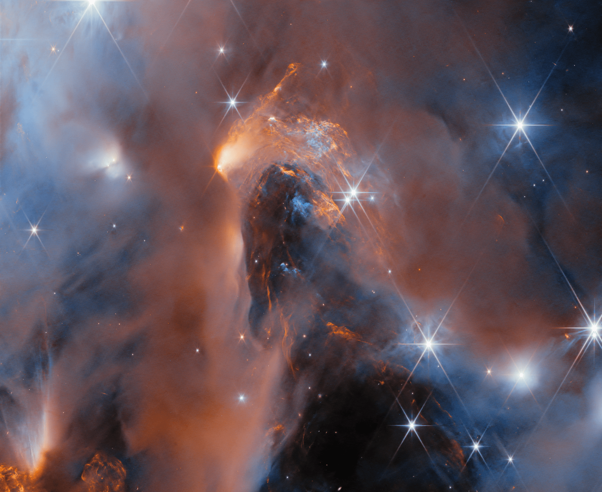 A nebula made up of cloudy gas and dust in the form of soft and wispy clouds and, in the center, thin and highly detailed layers pressed close together. Large, bright stars surrounded by six long points of light are dotted over the image, as well as some small, point-like stars embedded in the clouds. The clouds are lit up in blue close to the stars; orange colors show clouds that glow in infrared light.