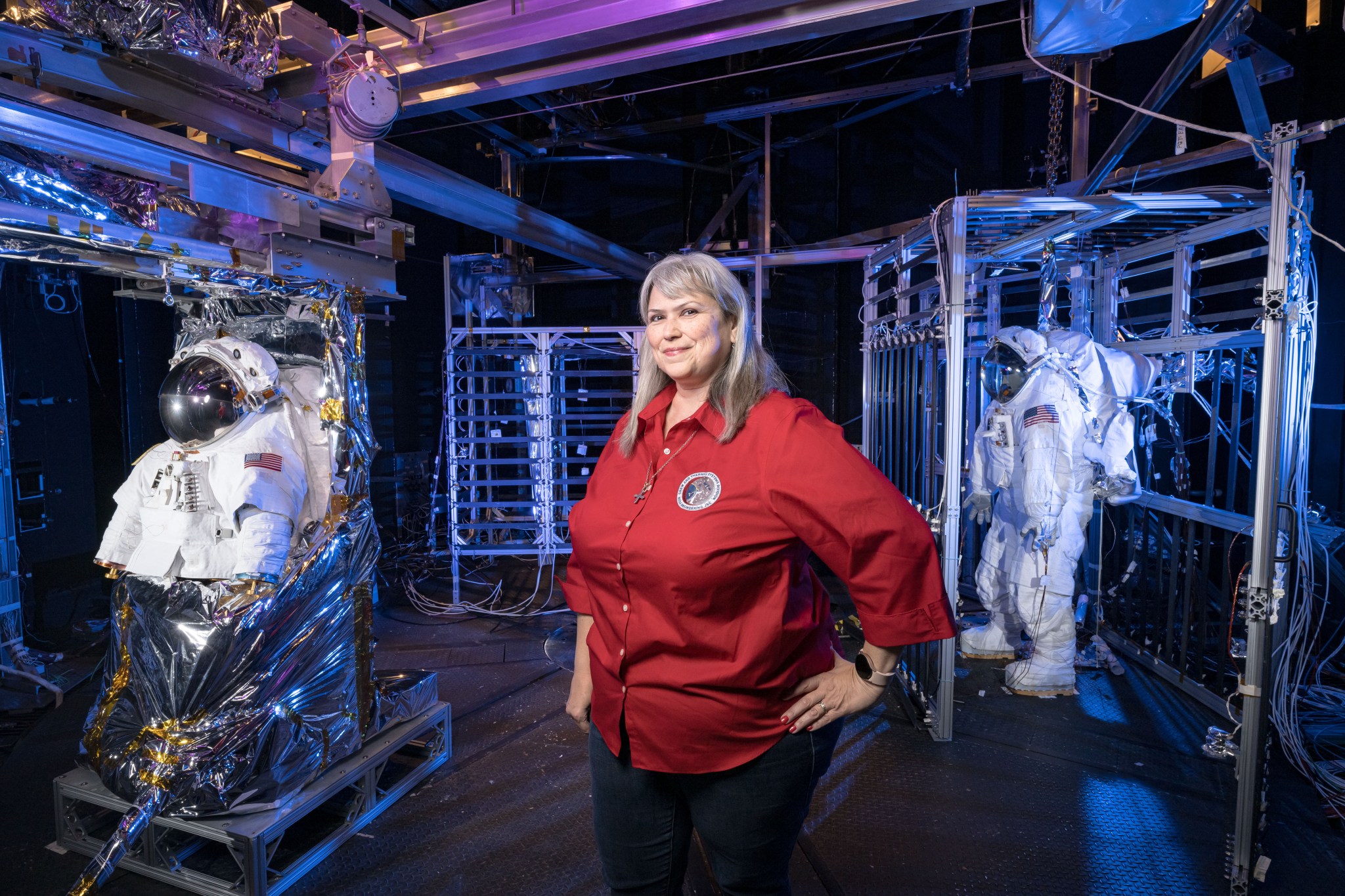 A NASA employee stands in a spacesuit test chamber with spacesuits in the background.