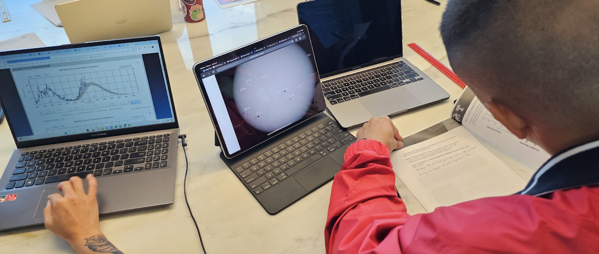 Two ambassadors work with NASA sunspot data on computers, graphing sunspot number versus time, and an image of the Sun's surface with spots, while completing an activity sheet.