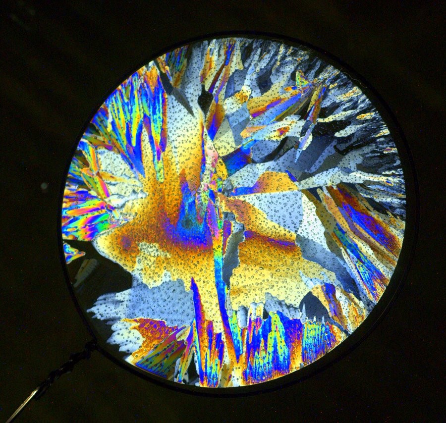 A circular piece of ice in front of a black background shows a jewel-like structure with a variety of yellows, blues, pinks, and greens. The ice has small gas bubbles spread throughout the structure with sharp breaks and jagged edges between the colors.