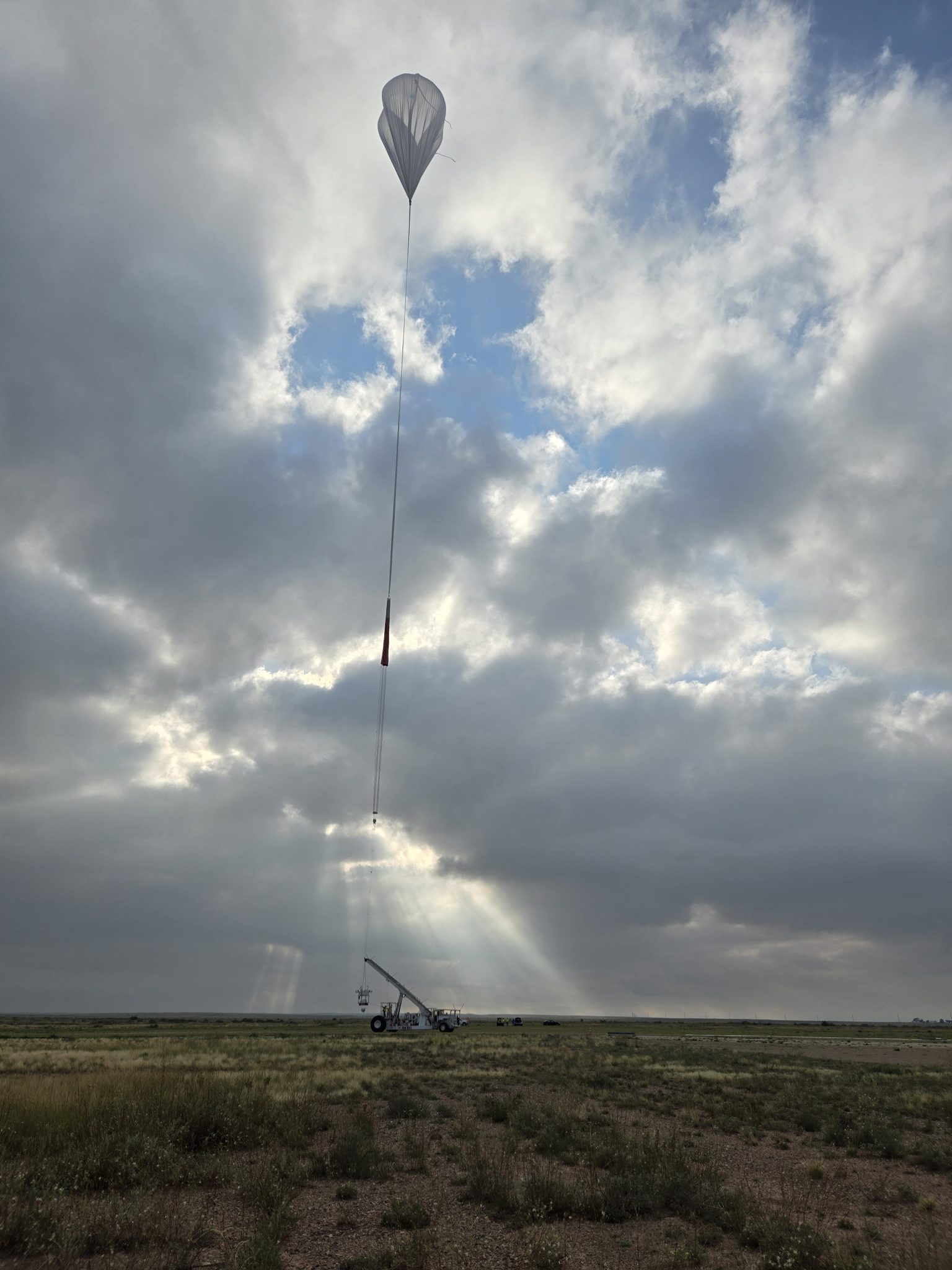 A NASA balloon lifts off connected to HASP payload attached to a launch vehicle. The Sun peeks through clouds, shining on the gondola.