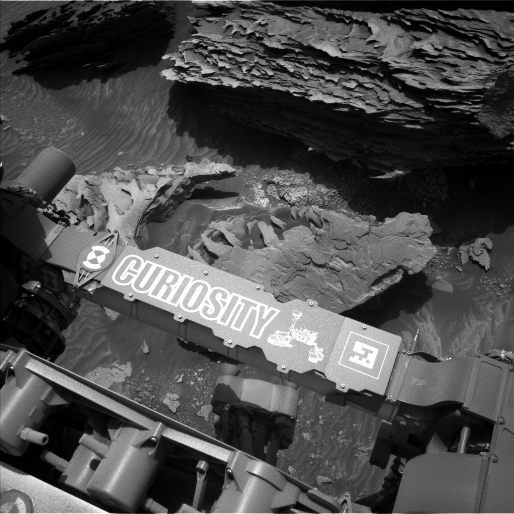 A grayscale photograph of the Martian surface from the Curiosity rover captures parts of the rover in the bottom half of the frame, including a crosspiece imprinted with its name and a line drawing of Curiosity. On the dark soil just below that is a very large, craggy rock in medium gray, while ahead of that is a much larger, much darker rock in the rover’s path, with the side showing looking like the raggedy edge view of the pages of a book torn in half.