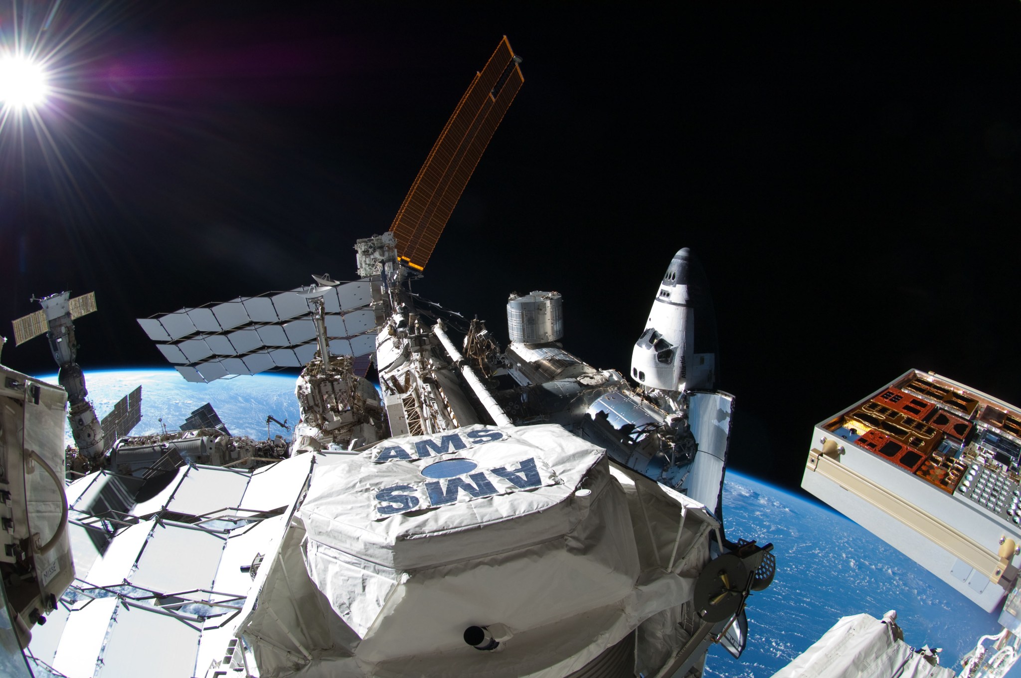 A white hexagonal-shaped cover over the top of the instrument is emblazoned with the letters “AMS” facing in two directions. There is a white cover over the large base of the instrument as well. Solar panels are visible to the left of and behind the instrument with another exterior instrument to its right. The blue curve of Earth is visible in the lower right, with black space above it. Bright rays stream from the Sun in the upper left corner.