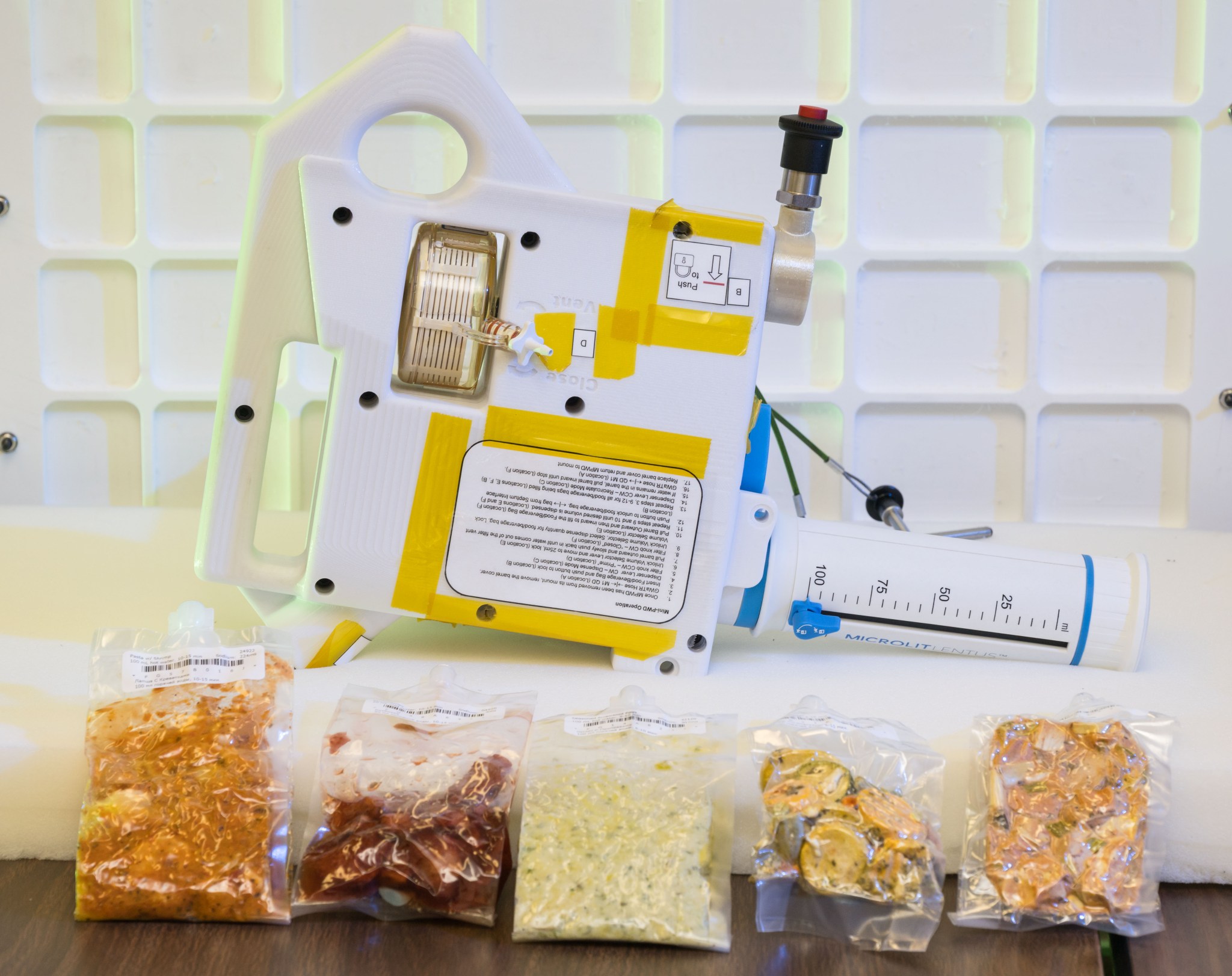 The image shows a compact, handheld water dispenser device placed on a table, with several vacuum-sealed packets of food arranged in front of it. The device is white with yellow tape and has various labels and instructions on its surface. It includes a large, graduated syringe-like component marked in milliliters, used for precise water measurement. The food packets in front of the device contain different types of rehydratable meals in various colors and textures, indicating the preparation of food for consumption in a space environment. The background features a white grid pattern, enhancing the focus on the device and food packets. This device is being developed for astronauts who live and work aboard Gateway.