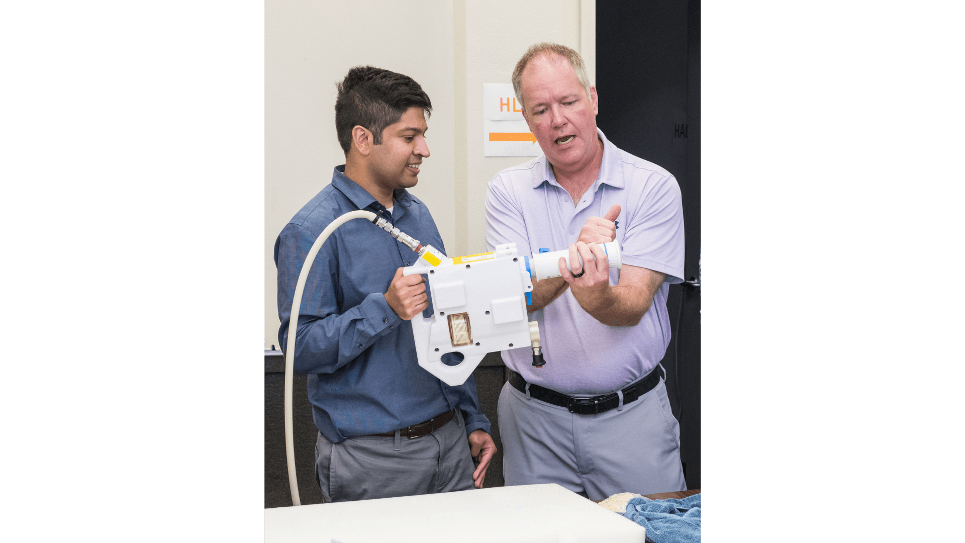 The image shows two individuals standing side by side as they demonstrate the Mini Potable Water Dispenser. The person on the left, dressed in a blue shirt, is holding the device, while the person on the right, wearing a light purple shirt, is explaining how to use it.