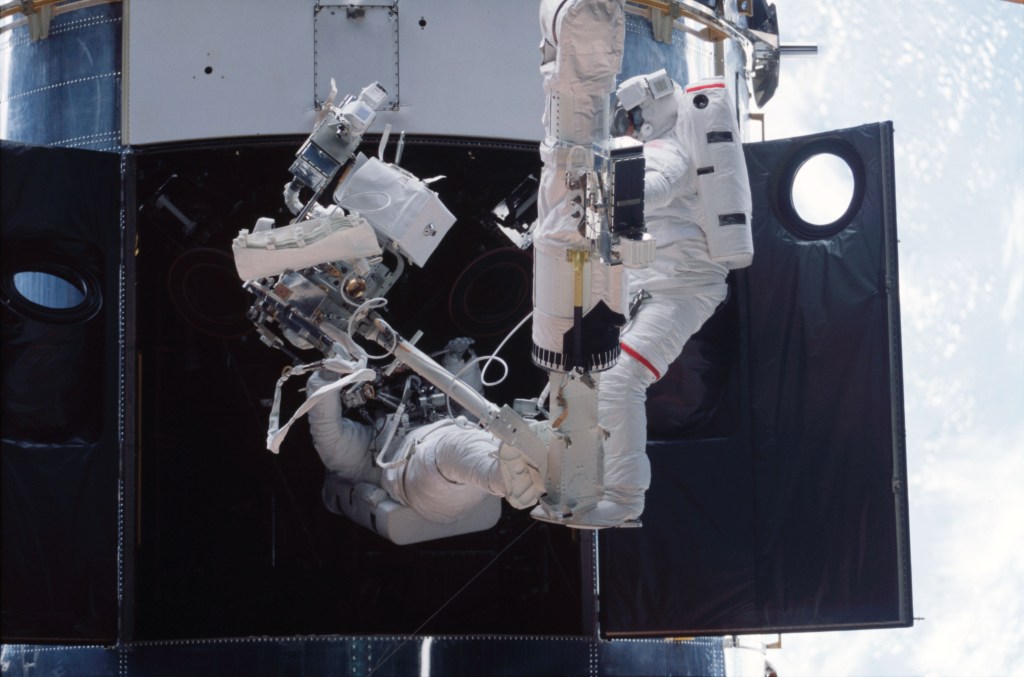 Two astronauts in spacesuits during a spacewalk working on the Hubble Space Telescope