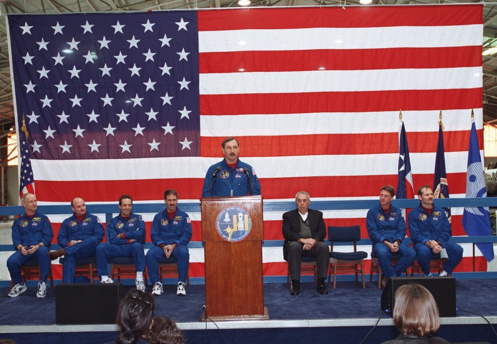 Seven astronauts seated on a stage, with one of them speaking into a microphone, with a very large US flag behind them