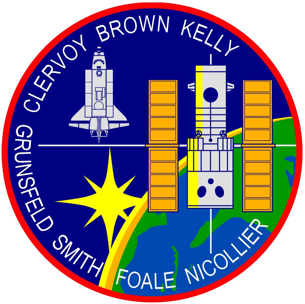 A circular crew patch, predominantly blue with a red rim, including a space shuttle, the Hubble Space Telescope, and the astronauts' names around the edge