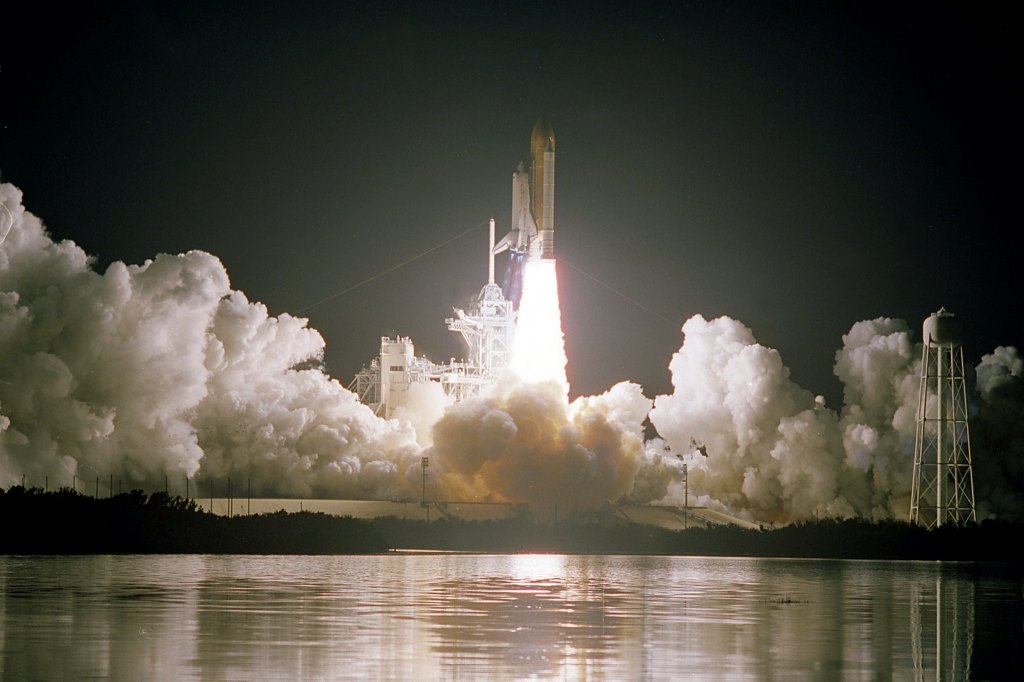 A space shuttle lifts off on a pillar of fire against a night sky