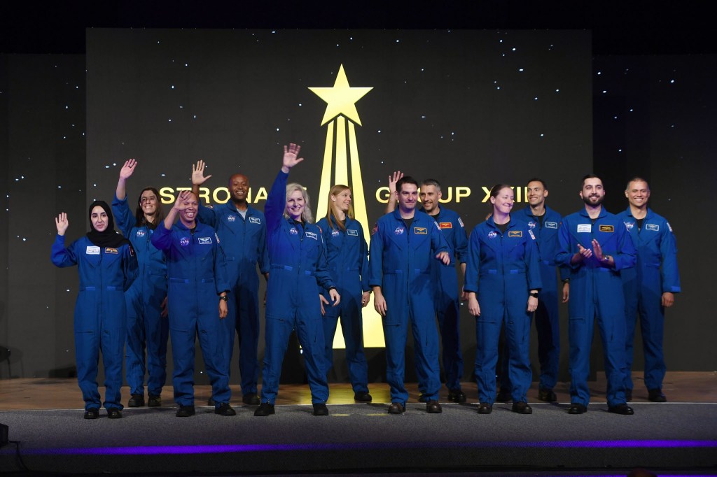 A group of new astronauts in their blue flight suits smile and wave from stage.