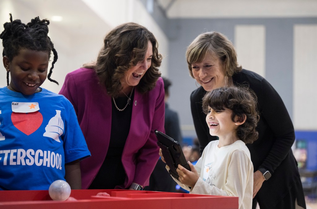 Two women are smiling and looking on as a beaming child operates a robot from a tablet.