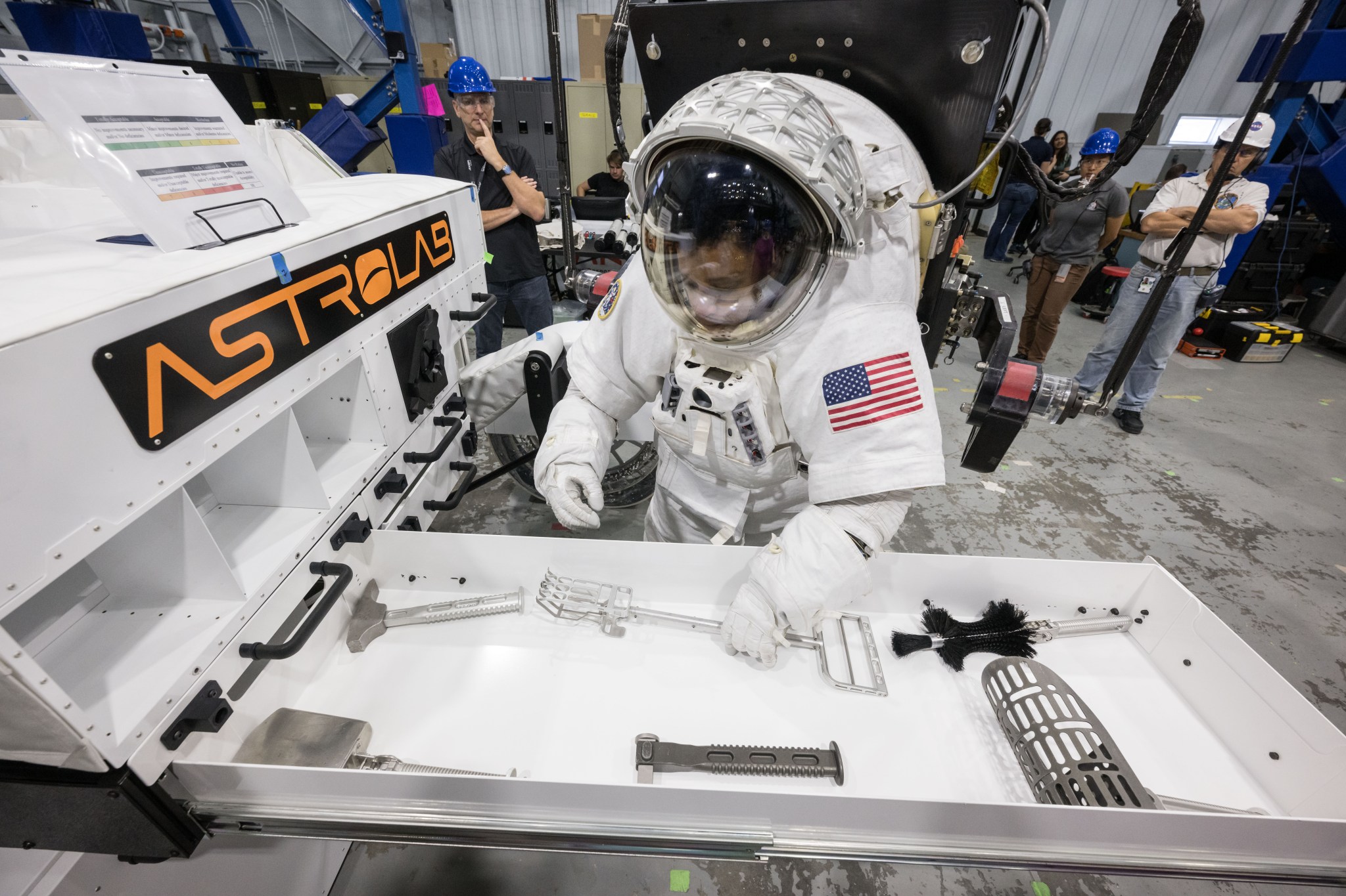 NASA astronaut Jessica Watkins is wearing a white spacesuit with the American flag on her left shoulder. She is pickup up a lunar geology tool from a tool drawer located outside of Astrolab's white lunar terrain vehicle. There is an orange Astrolab logo on the rover.