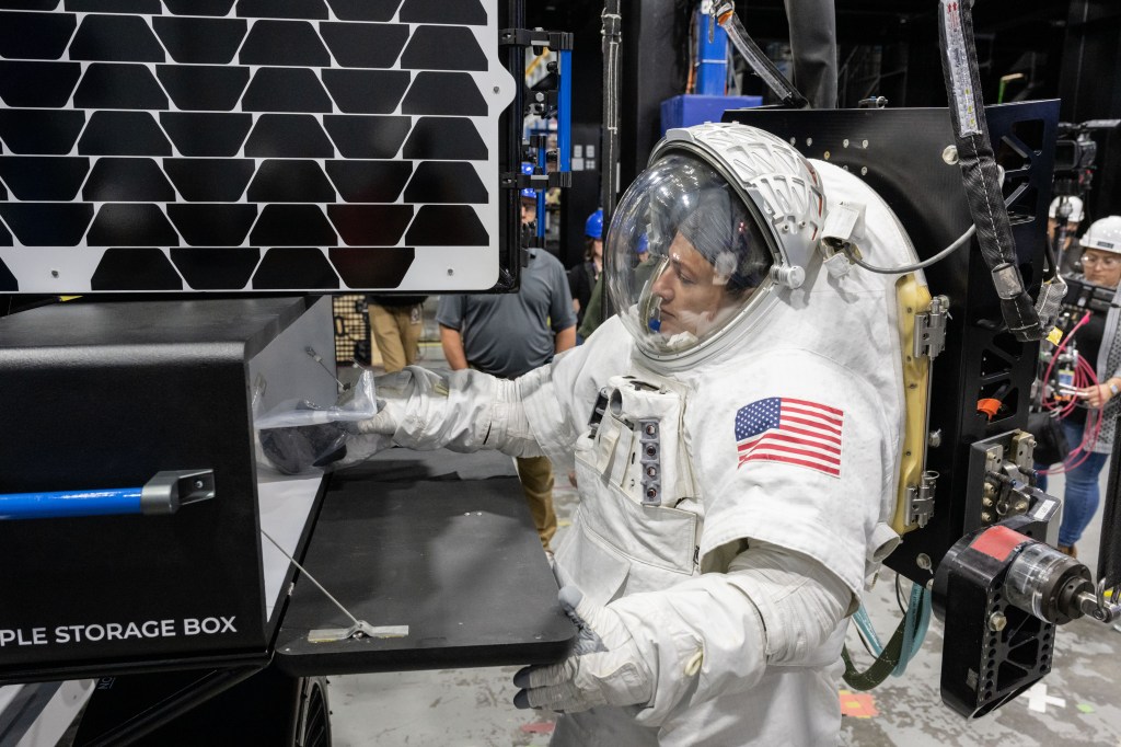 NASA astronaut Jessica Meir wearing a white spacesuit with the American flag on her left shoulder places a science sample bag inside of a black storage box with a blue handle.