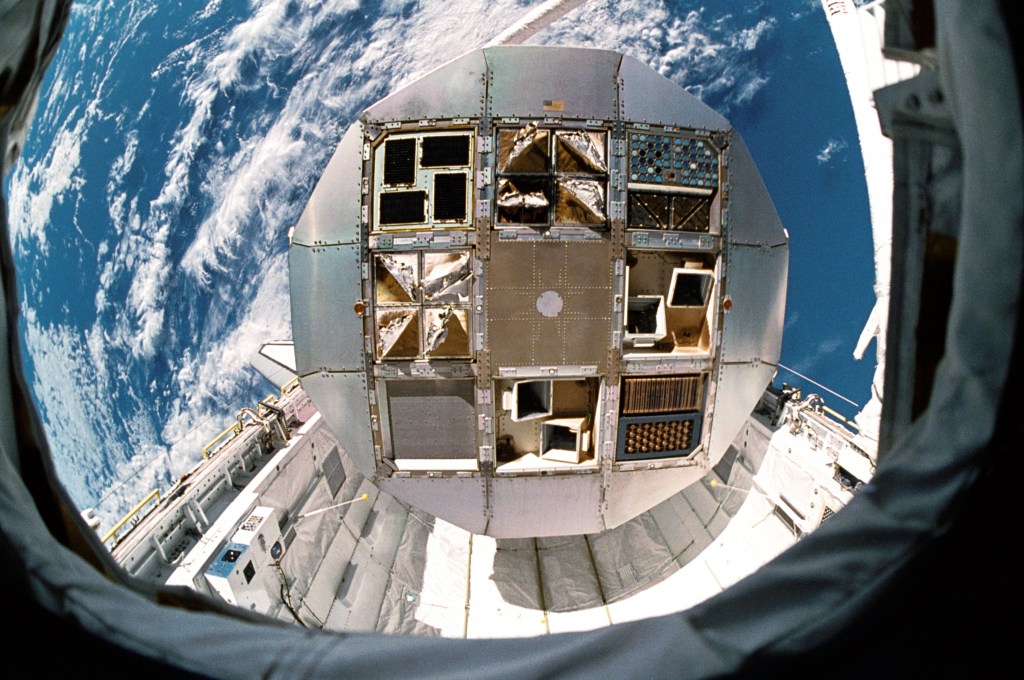 A large circular metal object, the end view of the satellite, seen against the blue and white Earth and the space shuttle payload bay