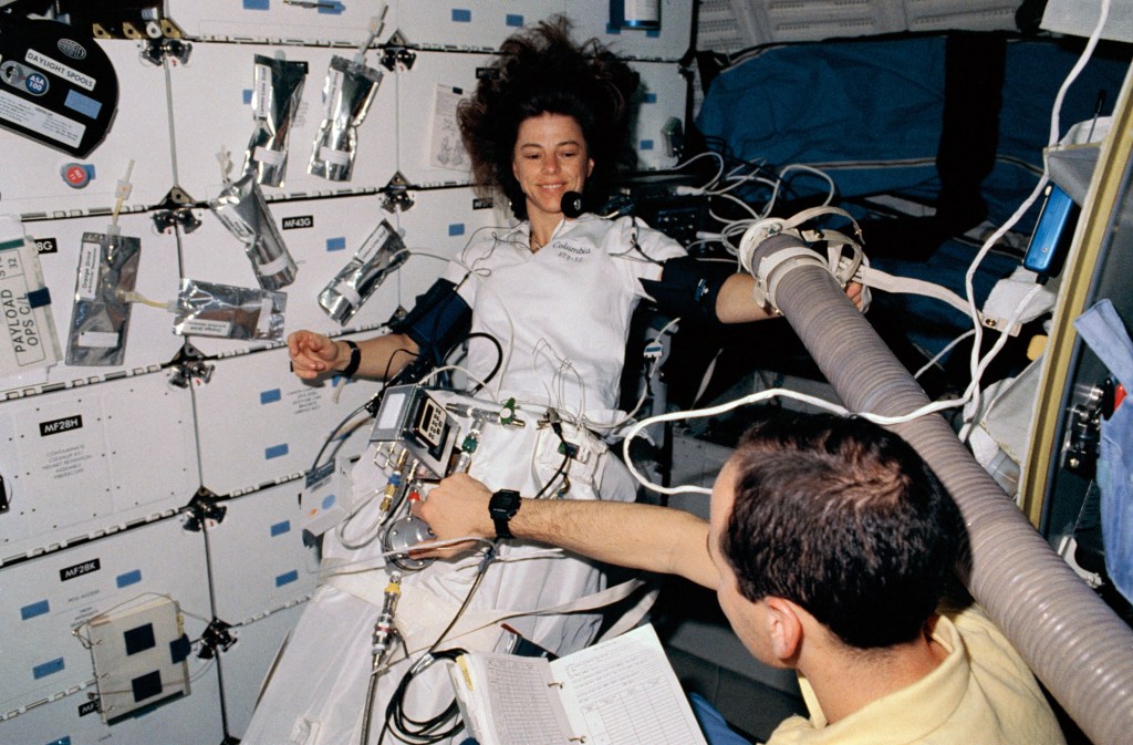 A view in the shuttle middeck, a woman astronaut is positioned inside a scientific equipment from the waist down while a male astronaut monitors the experiment.