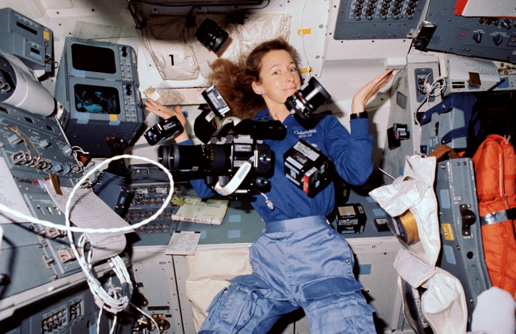 A woman astronaut floats in the shuttle with several cameras floating in front of her.