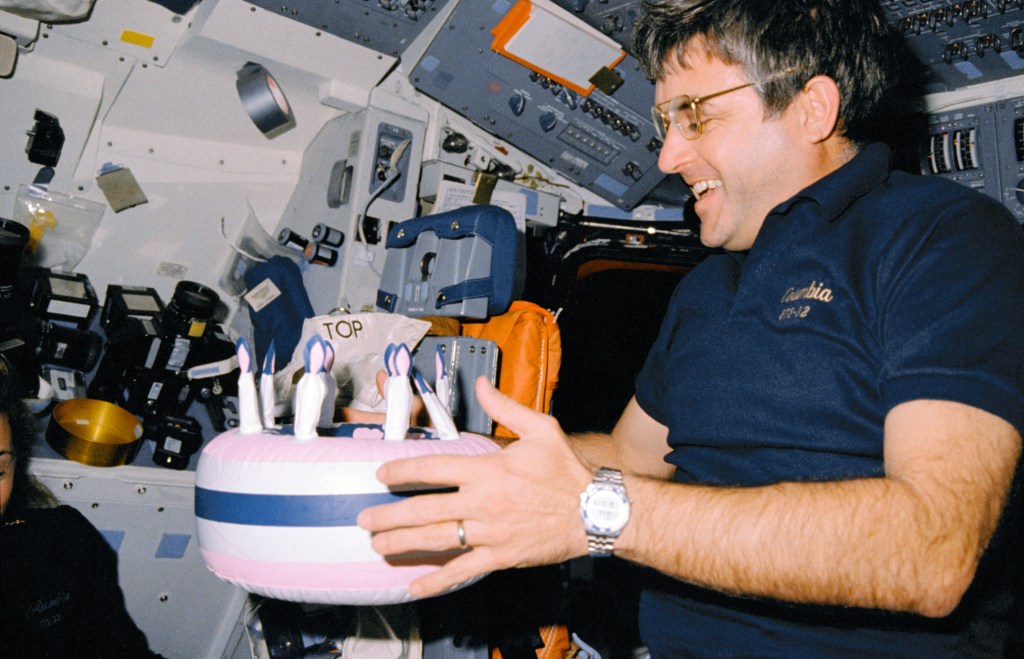 A male astronaut holds a plastic inflatable birthday cake while floating inside the space shuttle