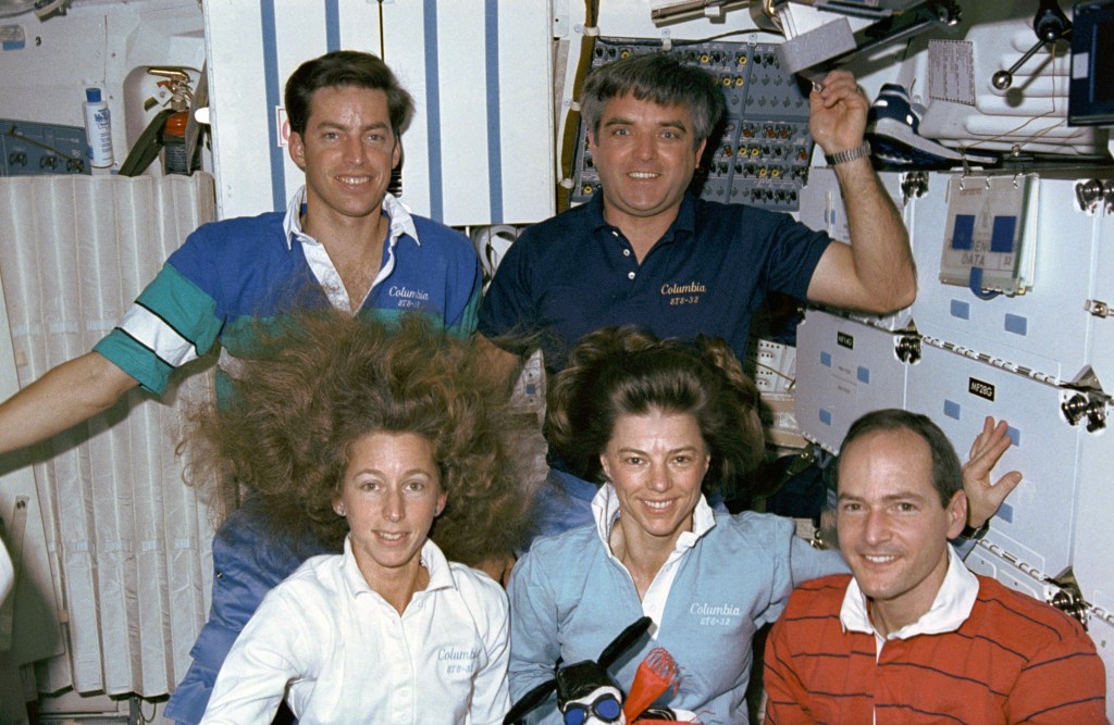 A group of three men and two women pose as a group, three in front, two in back, while floating inside the space shuttle.